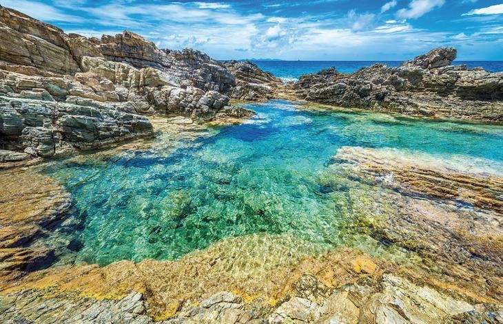 Crystal-clear turquoise water fills a natural rock pool surrounded by rugged coastal rocks under a bright blue sky.