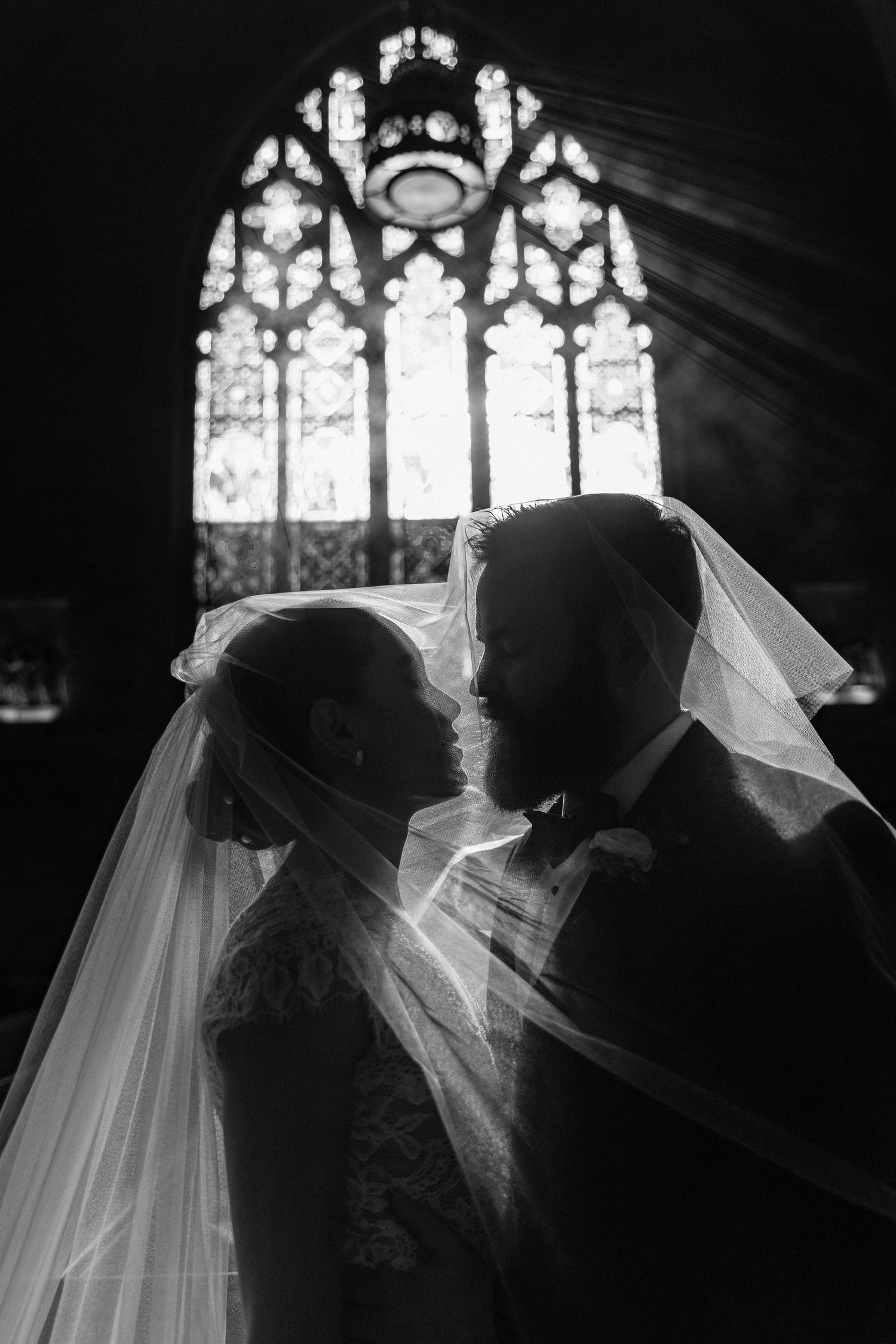 Black and white photo of bride and groom sharing intimate moment under veil before stained glass church window.