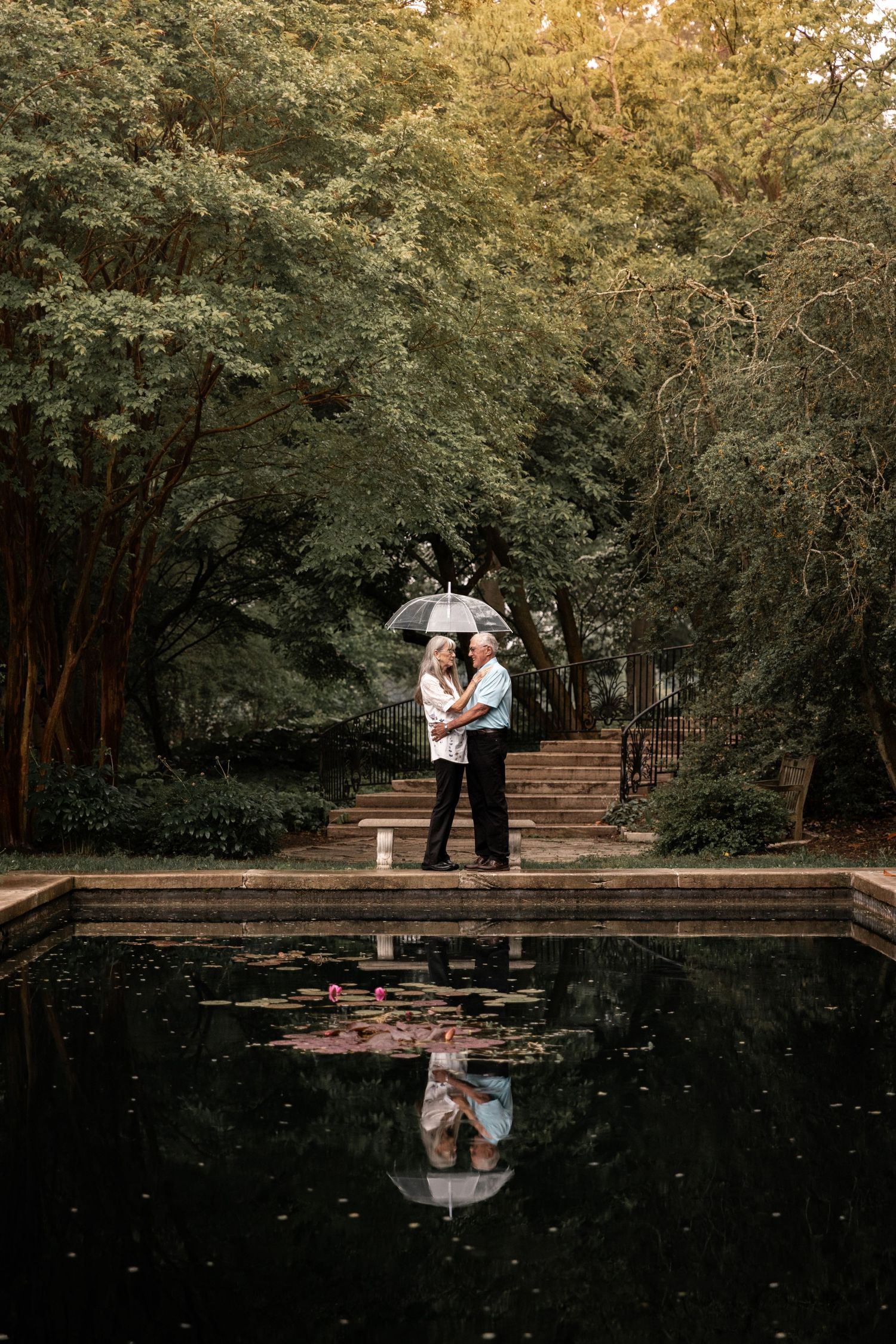 Couple sharing intimate moment with umbrella by reflecting pool surrounded by lush greenery.