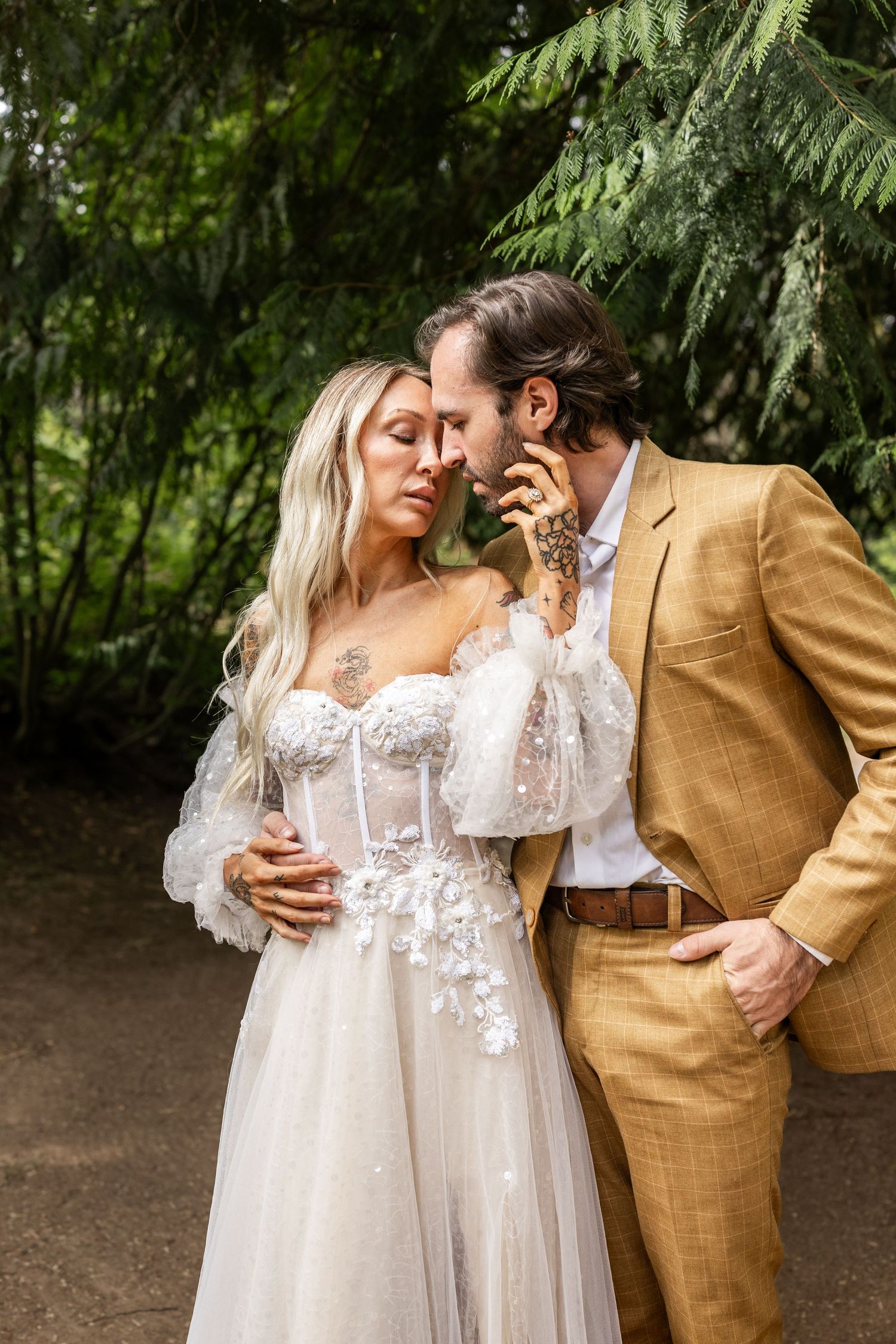 A bride in a white floral corset gown and groom in a tan suit share an intimate moment in a lush garden setting.