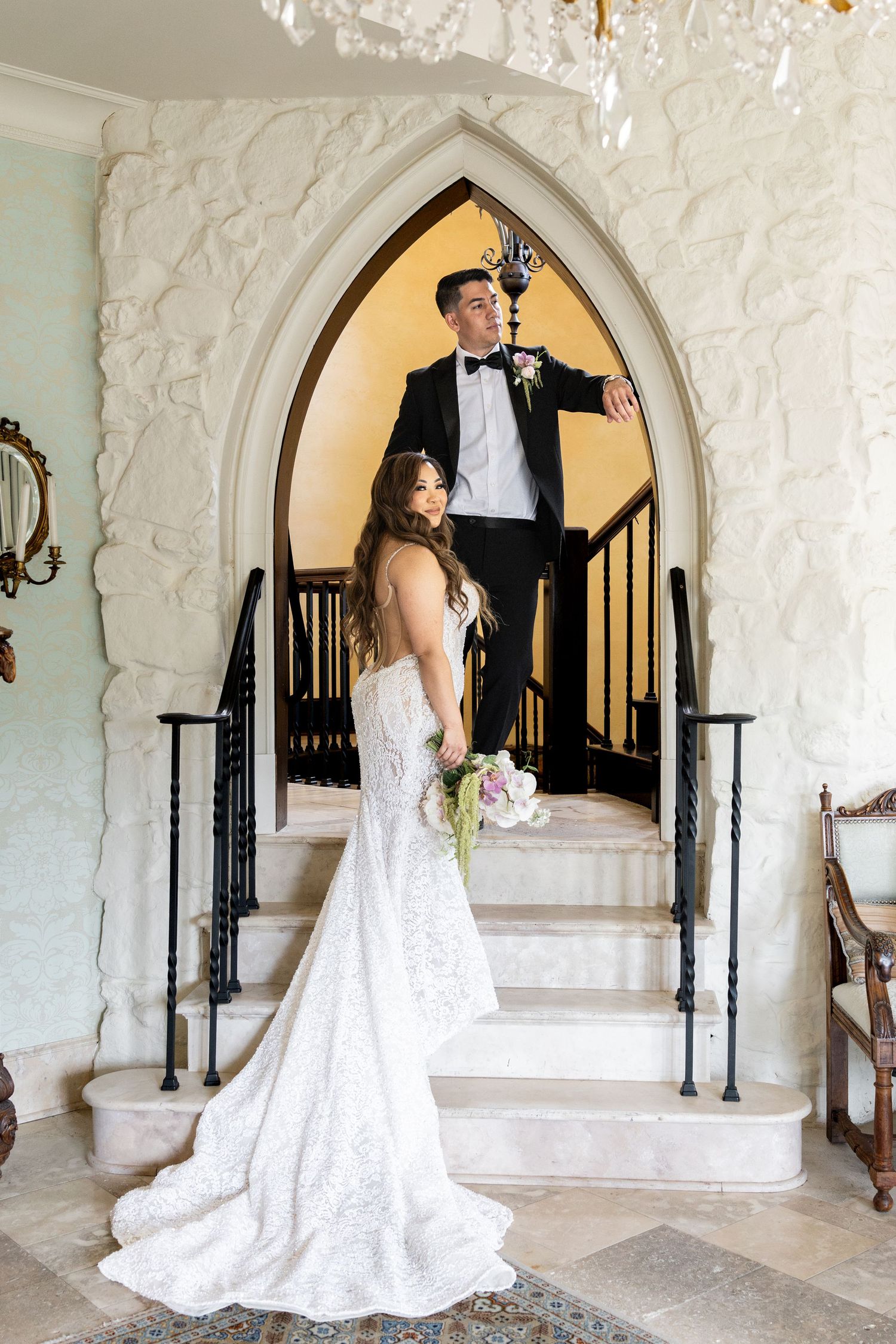 Bride in lace gown holds bouquet as groom in tuxedo stands on elegant staircase beneath Gothic arch in white venue.