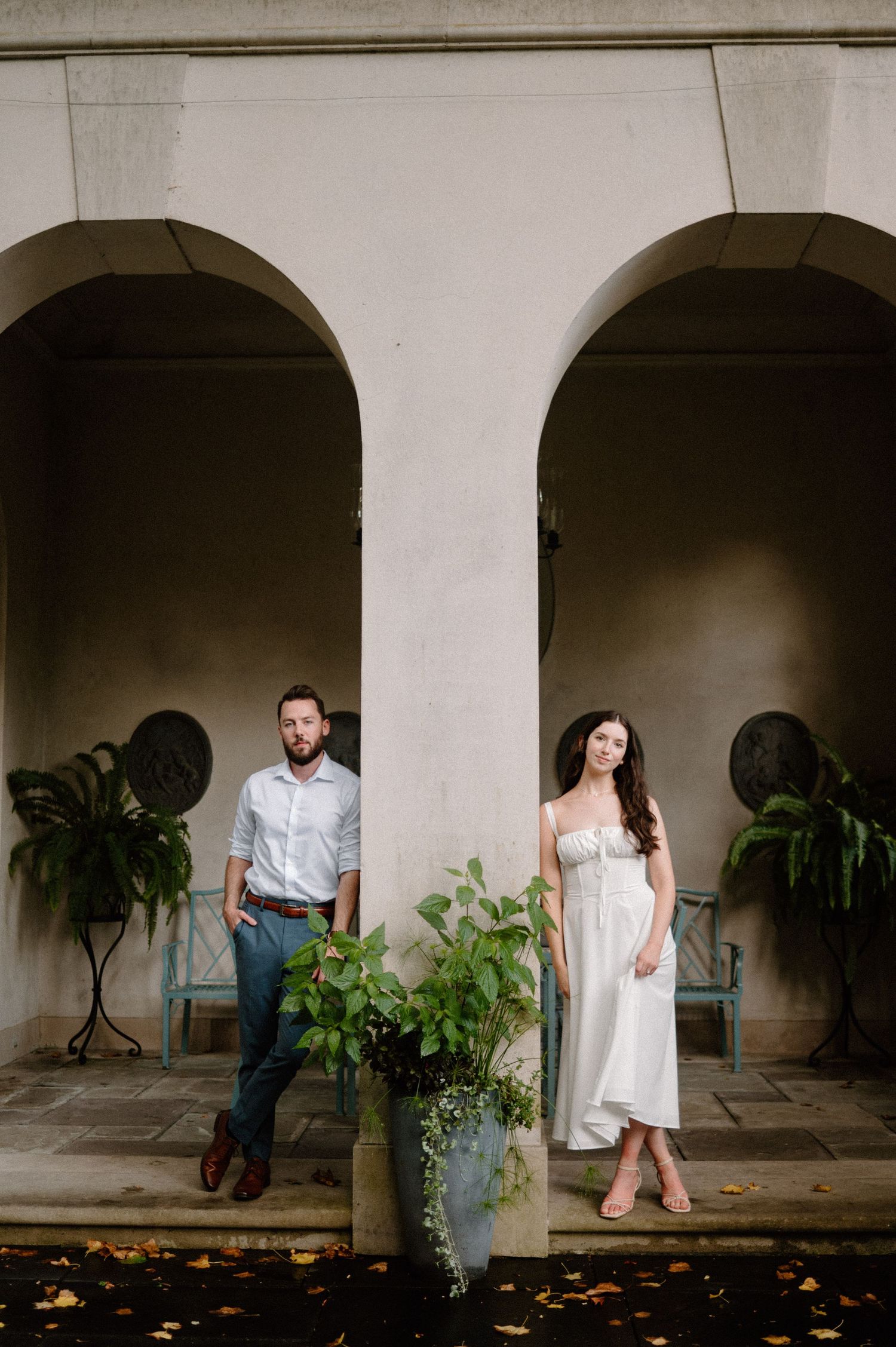 Stylish couple standing in separate stone archways with potted plants and white walls at an elegant venue.