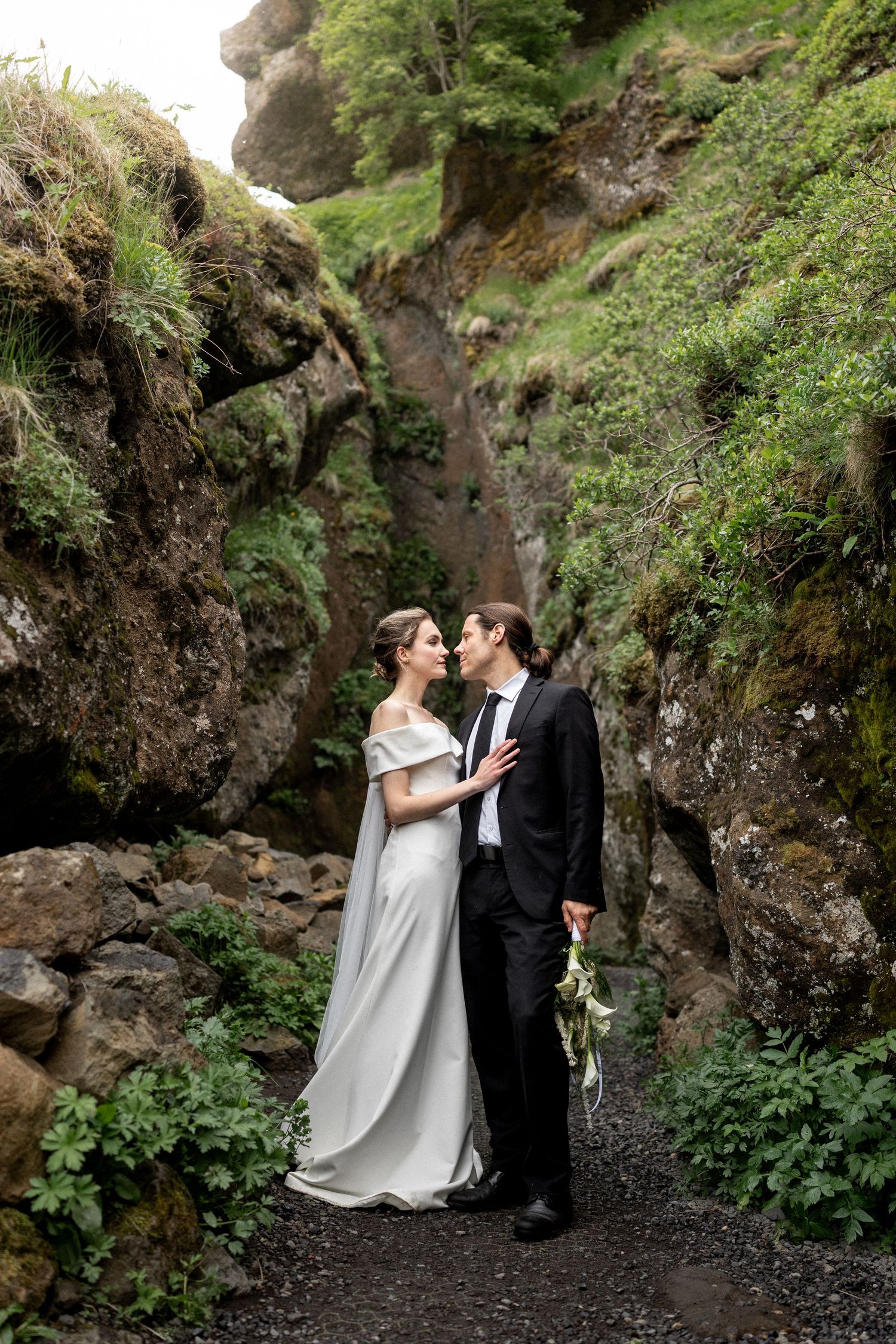 Bride and groom share a romantic moment in a lush green canyon, bride in white gown, groom in black suit holding bouquet.