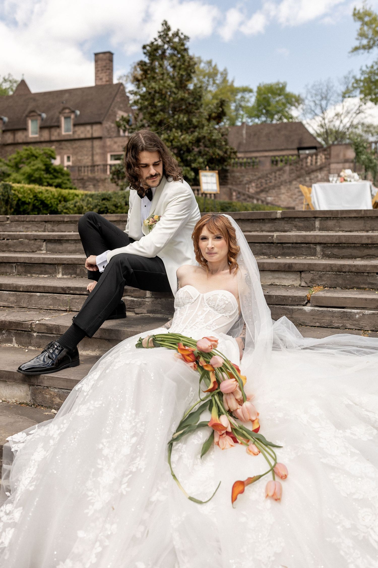 Bride in white lace gown with tulip bouquet sits on steps with groom in white tuxedo jacket outdoors.