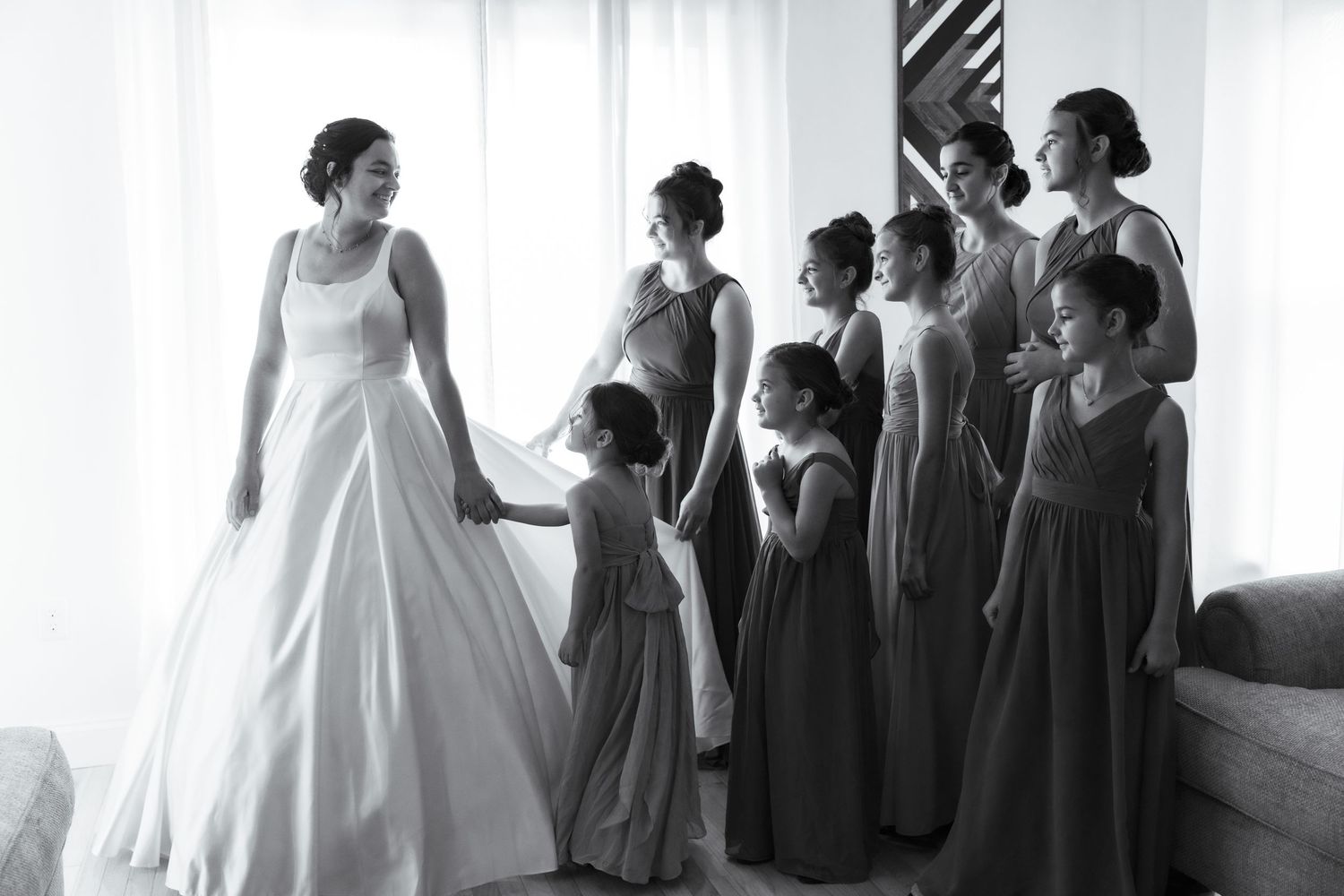 A bride and bridesmaids in gray dresses prepare for a wedding ceremony in a bright white room.