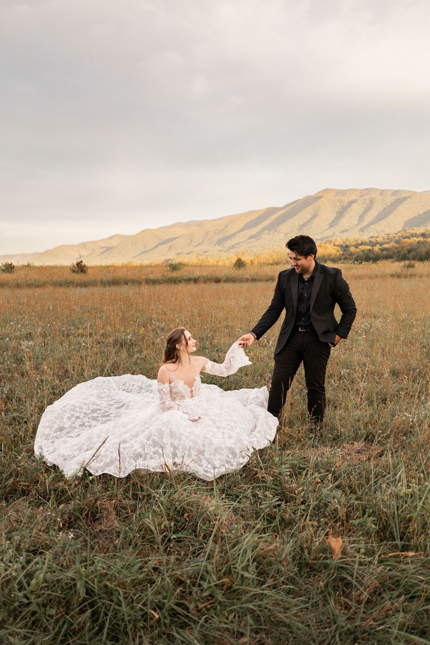 Bride in white gown sits in field as groom in black suit holds her hand, mountains in background.
