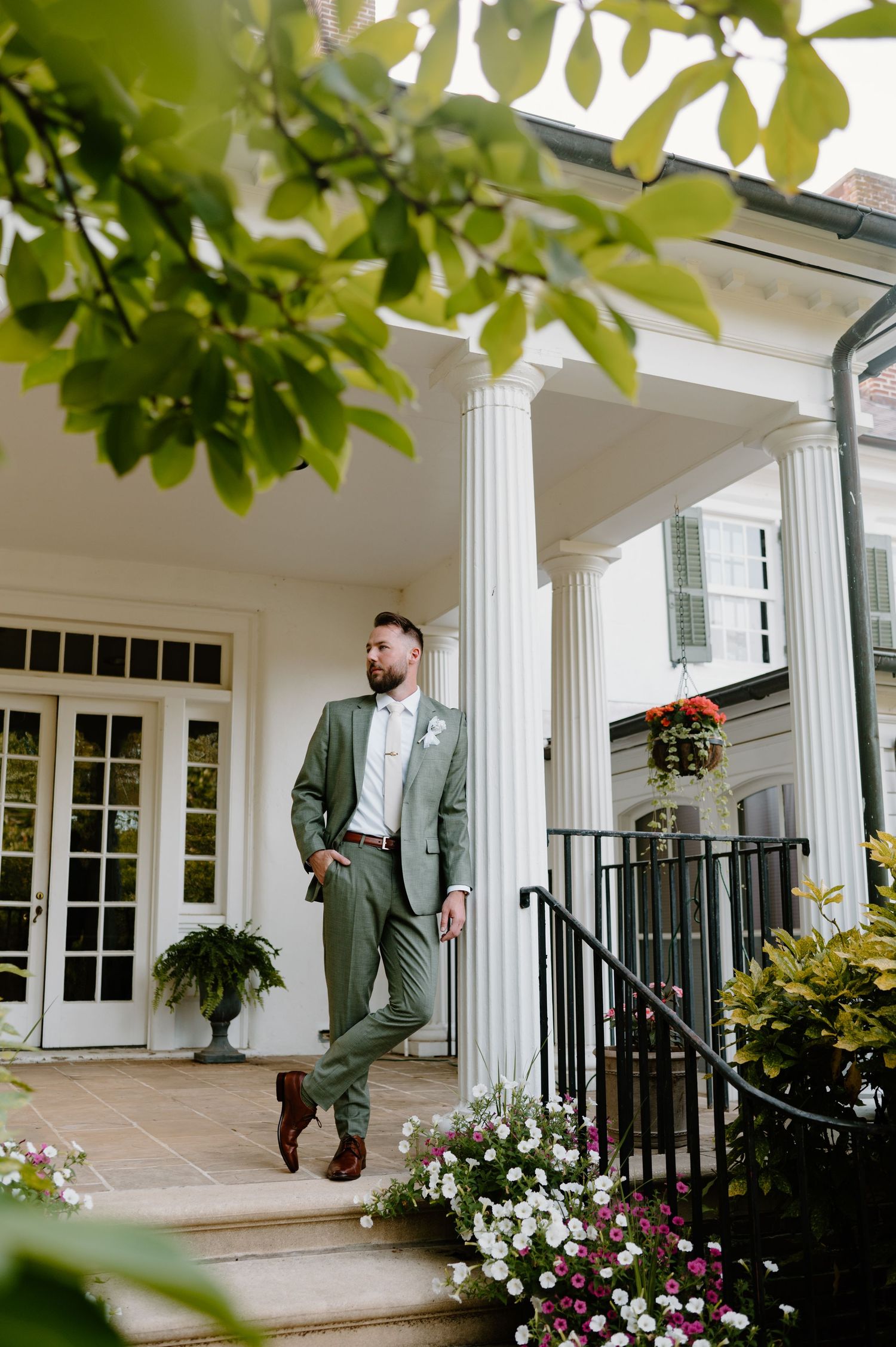 Groom in sage green suit stands on elegant white columned porch surrounded by lush greenery and colorful flowers.