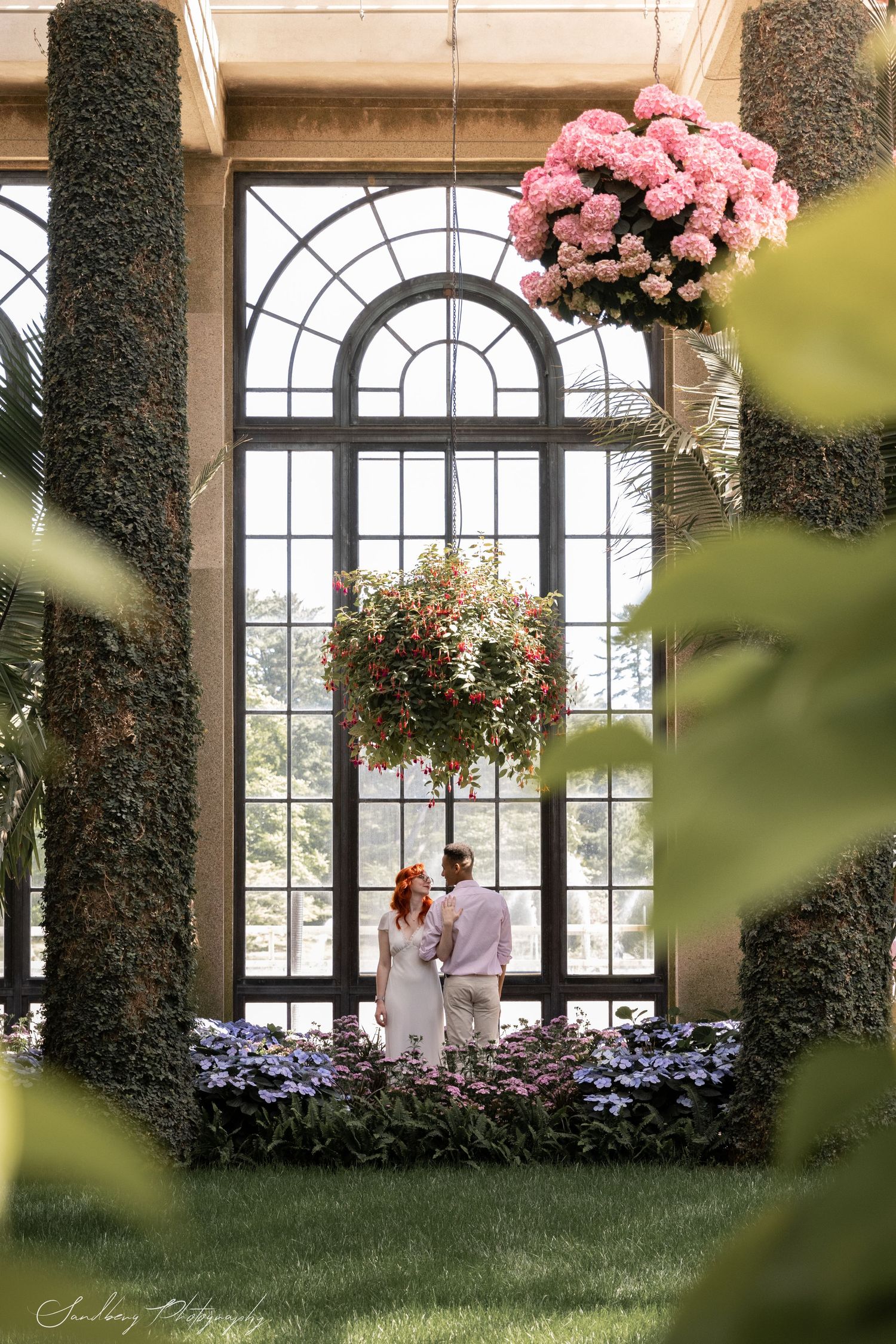 Couple in white attire beneath grand arched window with hanging pink floral arrangements.