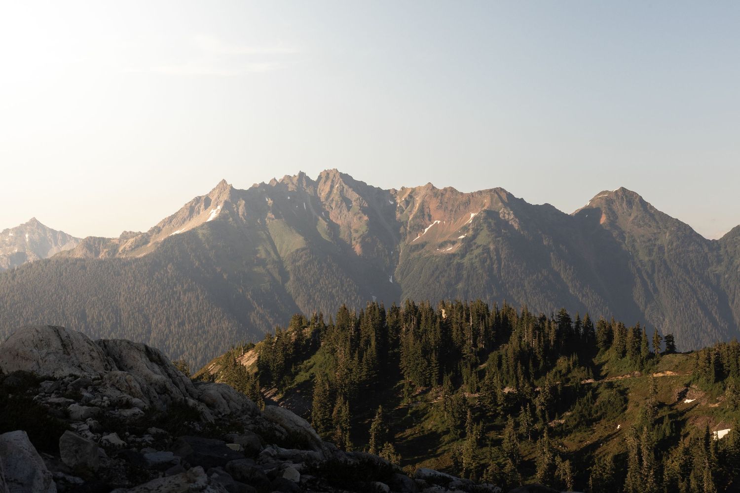 Panoramic mountain landscape with jagged snow-capped peaks and evergreen forest under cloudy skies at sunset.