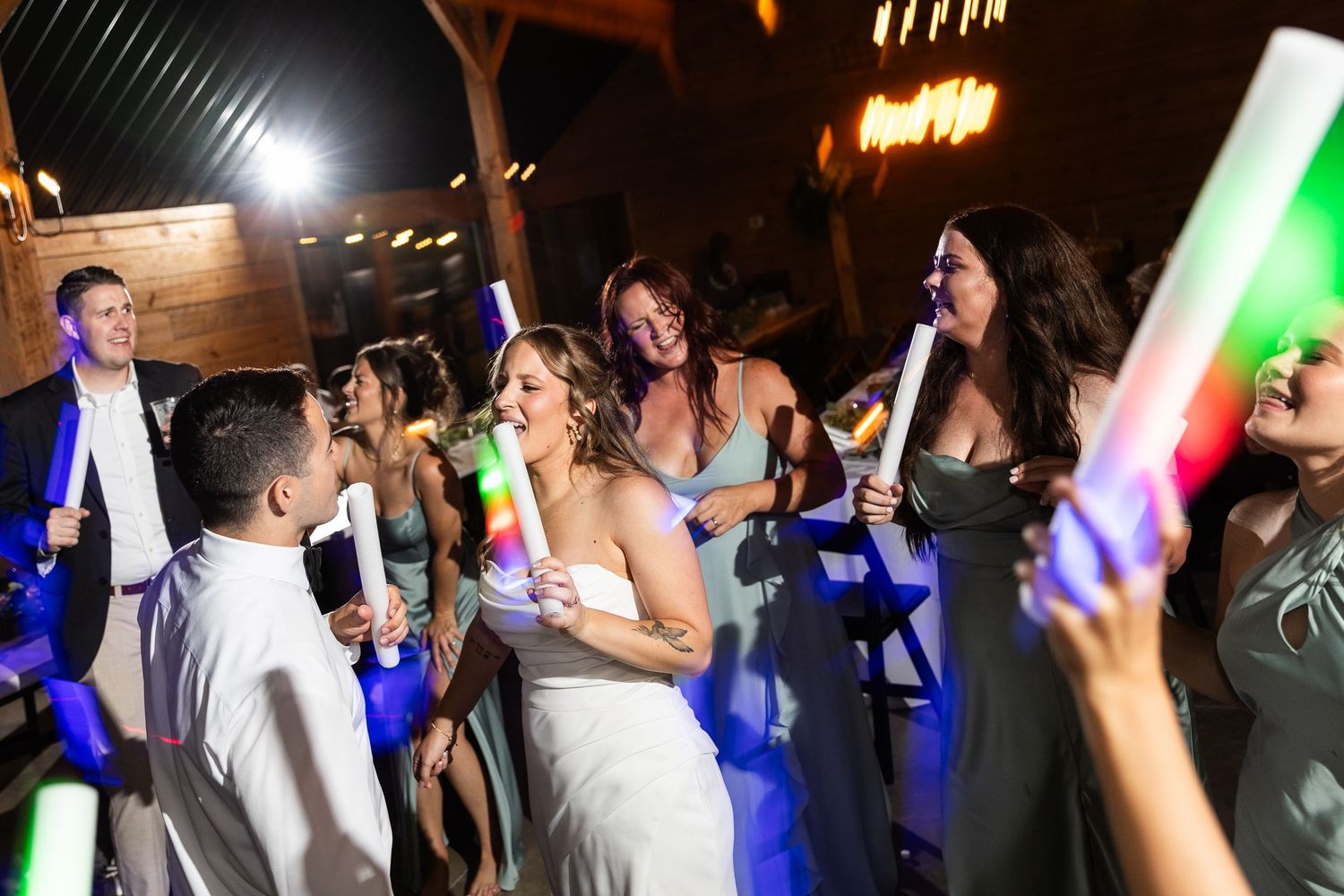 Wedding guests dance and celebrate on a dark dance floor illuminated by glowing LED foam sticks and colored lights.
