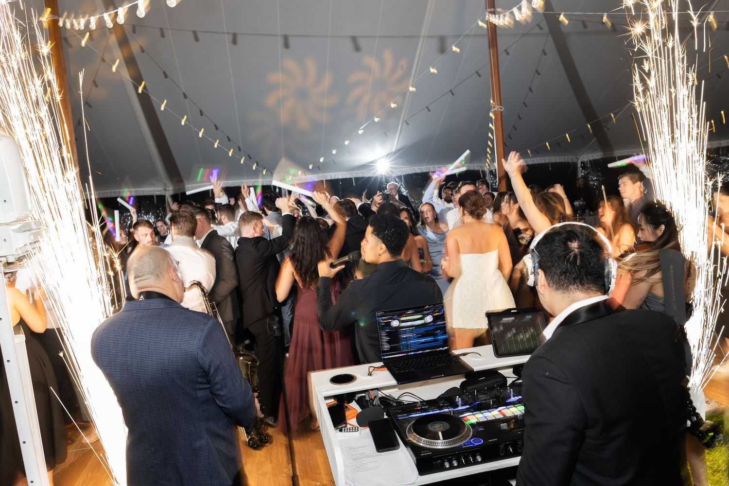 Wedding reception guests dance and celebrate under a tent while a DJ performs with sparkling lights illuminating the space.