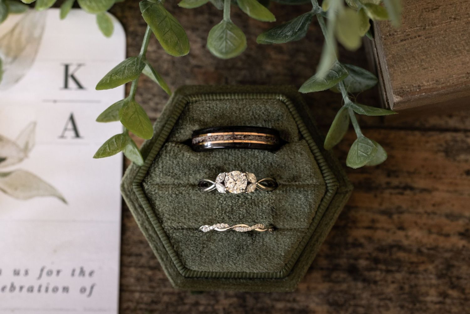 Three wedding rings displayed in a dark green velvet hexagonal ring box with eucalyptus leaves and invitation visible.