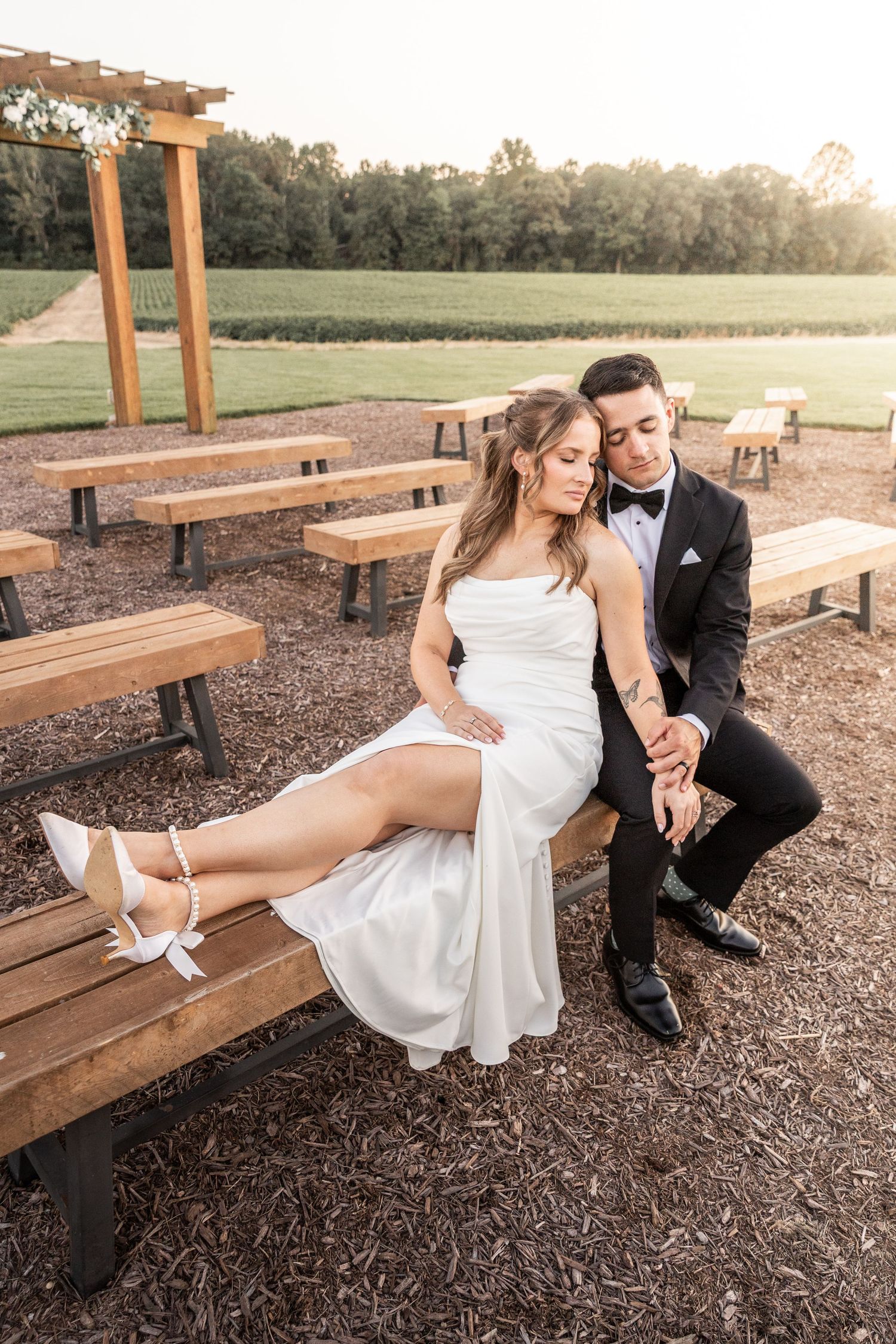 A romantic couple in wedding attire share tender moments on wooden benches at sunset in a rustic outdoor setting.
