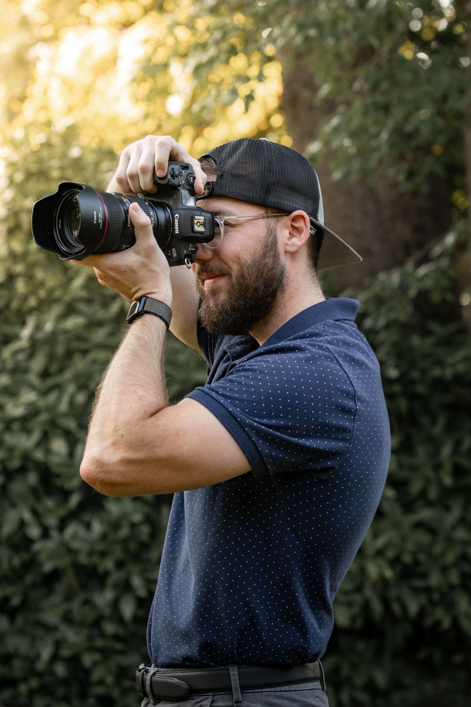 Bearded man in blue polo and cap photographing with a professional DSLR camera outdoors in natural green setting.