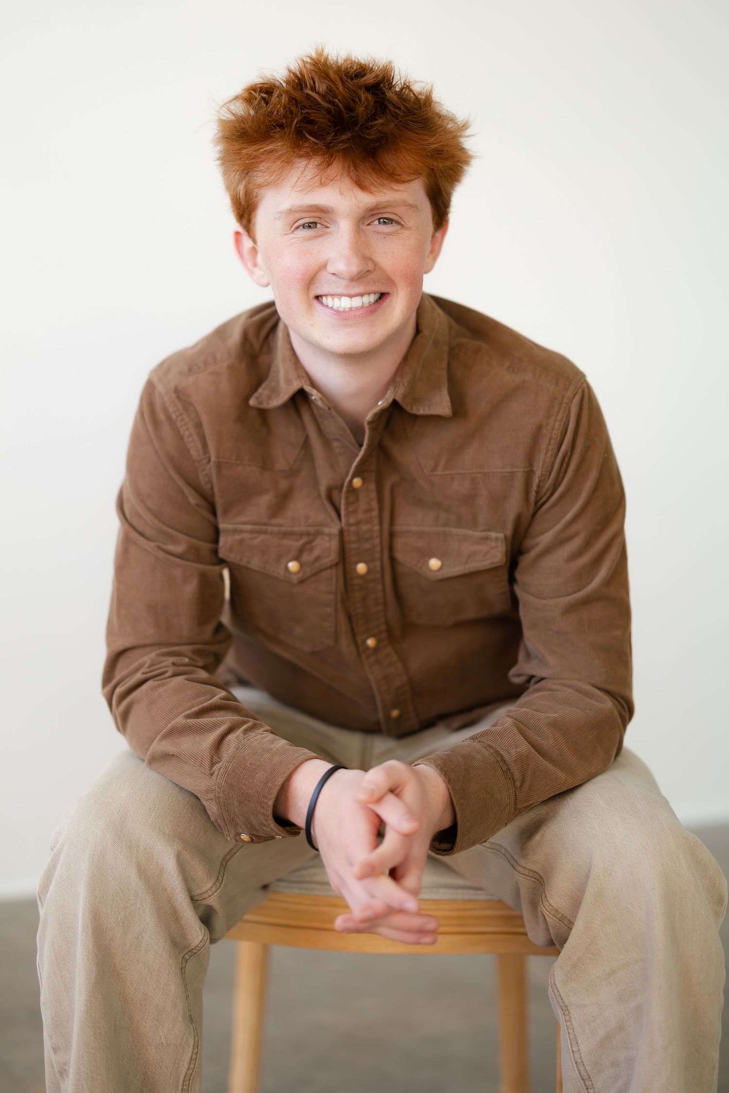 A person in a brown button-up shirt and khaki pants sits casually on a wooden stool against a light background.