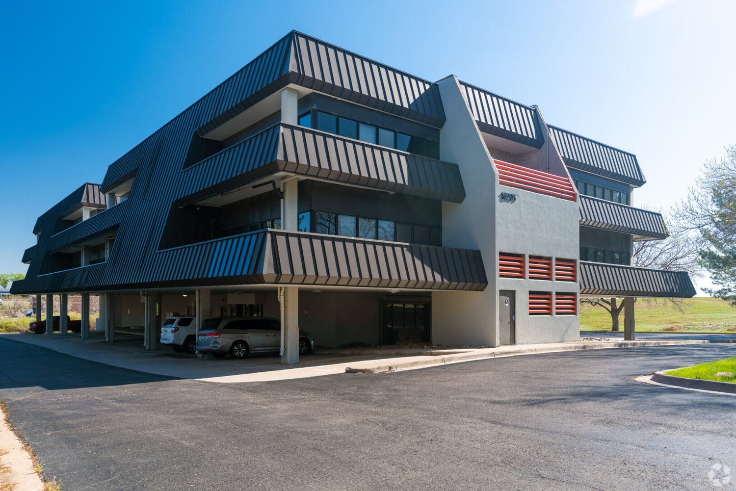 Modern multi-story commercial building with covered parking, metal facade, and balconies under a clear blue sky.