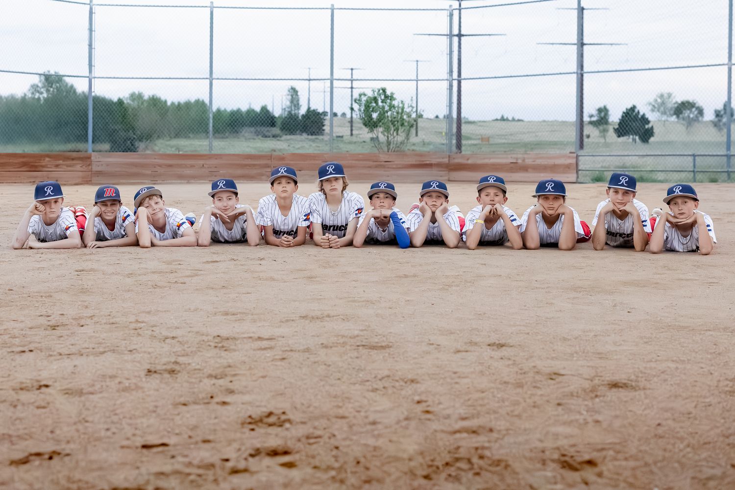 Young baseball team in uniforms lying on dirt field posing for team photo at outdoor baseball diamond.