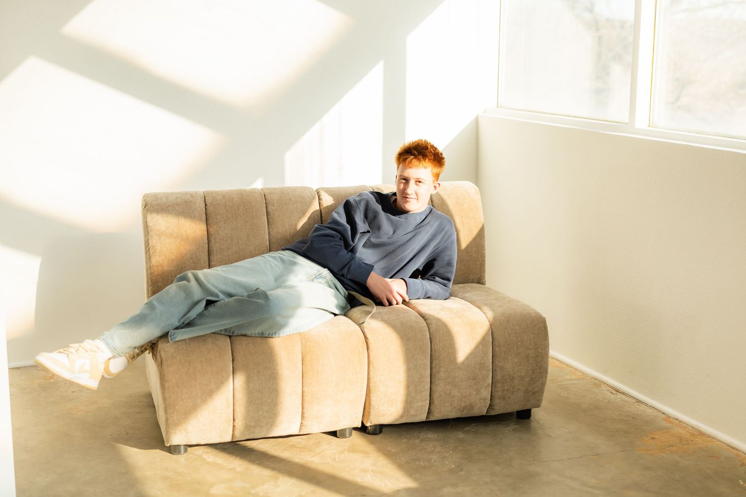 8th grade boy lies comfortably on a white couch in a sunlit room with minimalist decor.