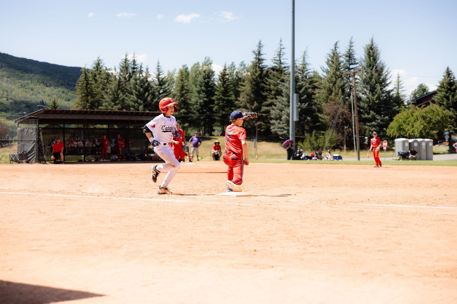 Youth baseball players running bases on dirt field with trees and mountains visible in background on sunny day.