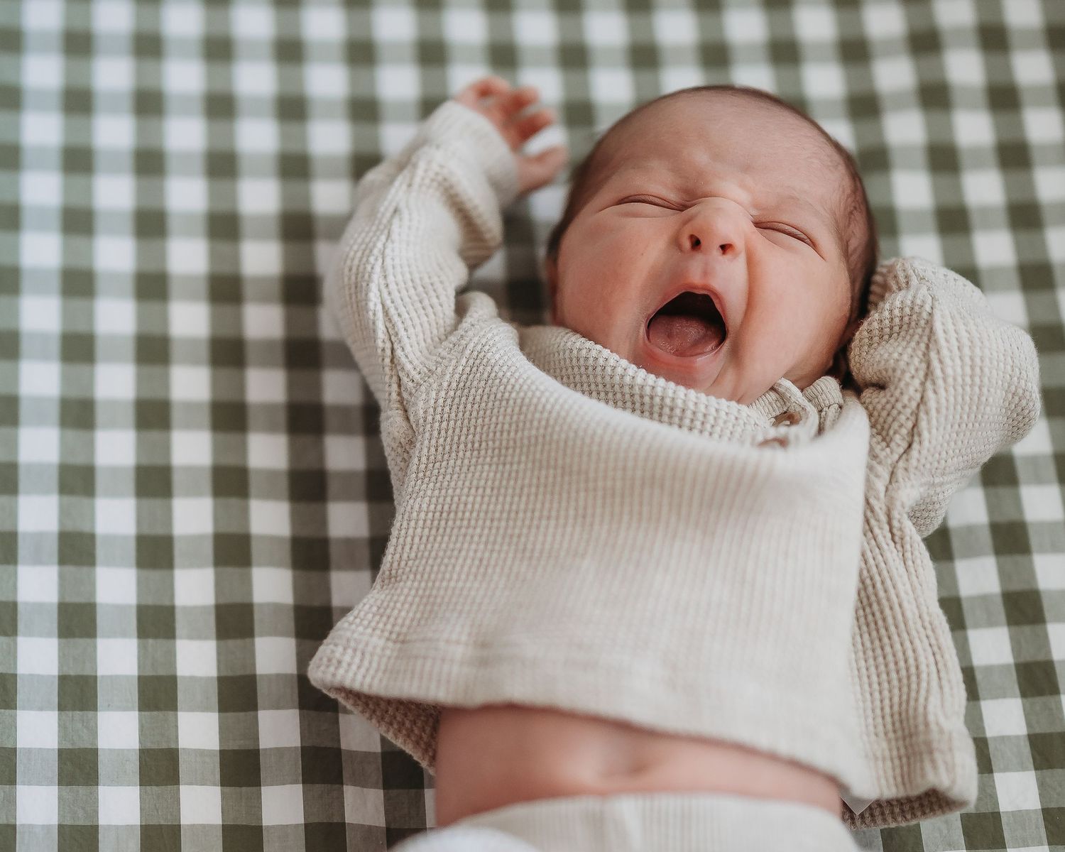 A baby in a cream sweater yawning while lying on a green checkered blanket.