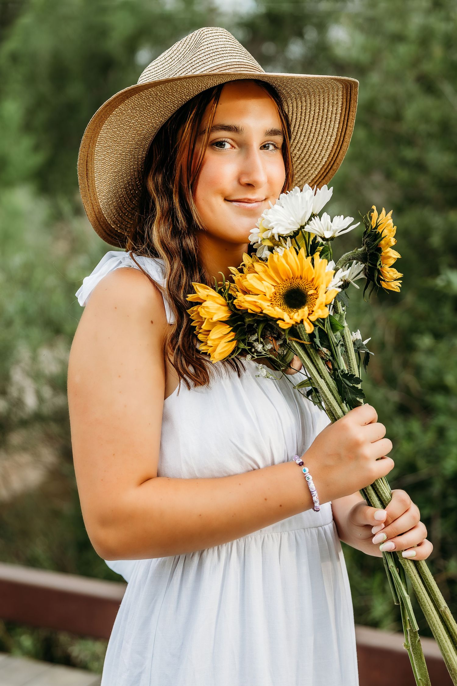 Person in white dress and sun hat holding bright yellow sunflowers in an outdoor garden setting.