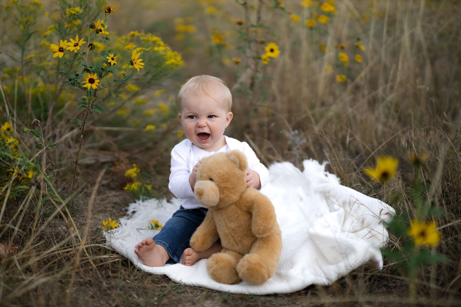 Baby on white blanket with teddy bear surrounded by yellow wildflowers.