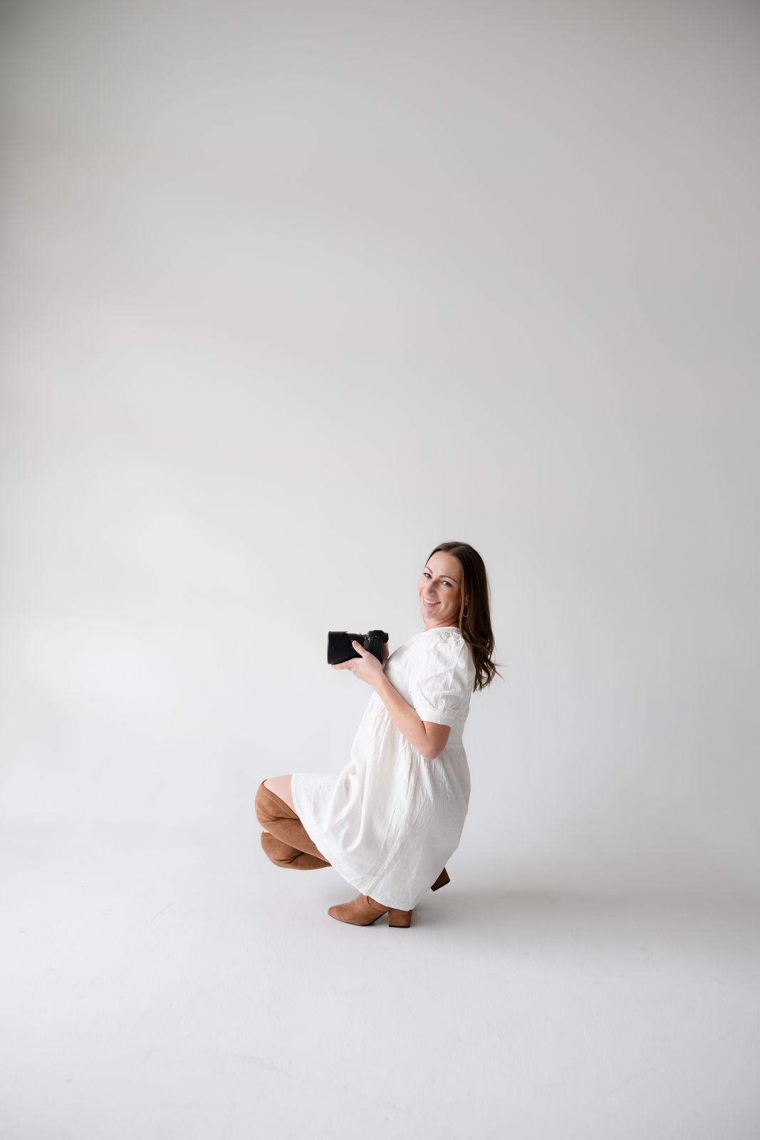 Photographer in white dress crouches with camera in minimalist white studio setting.
