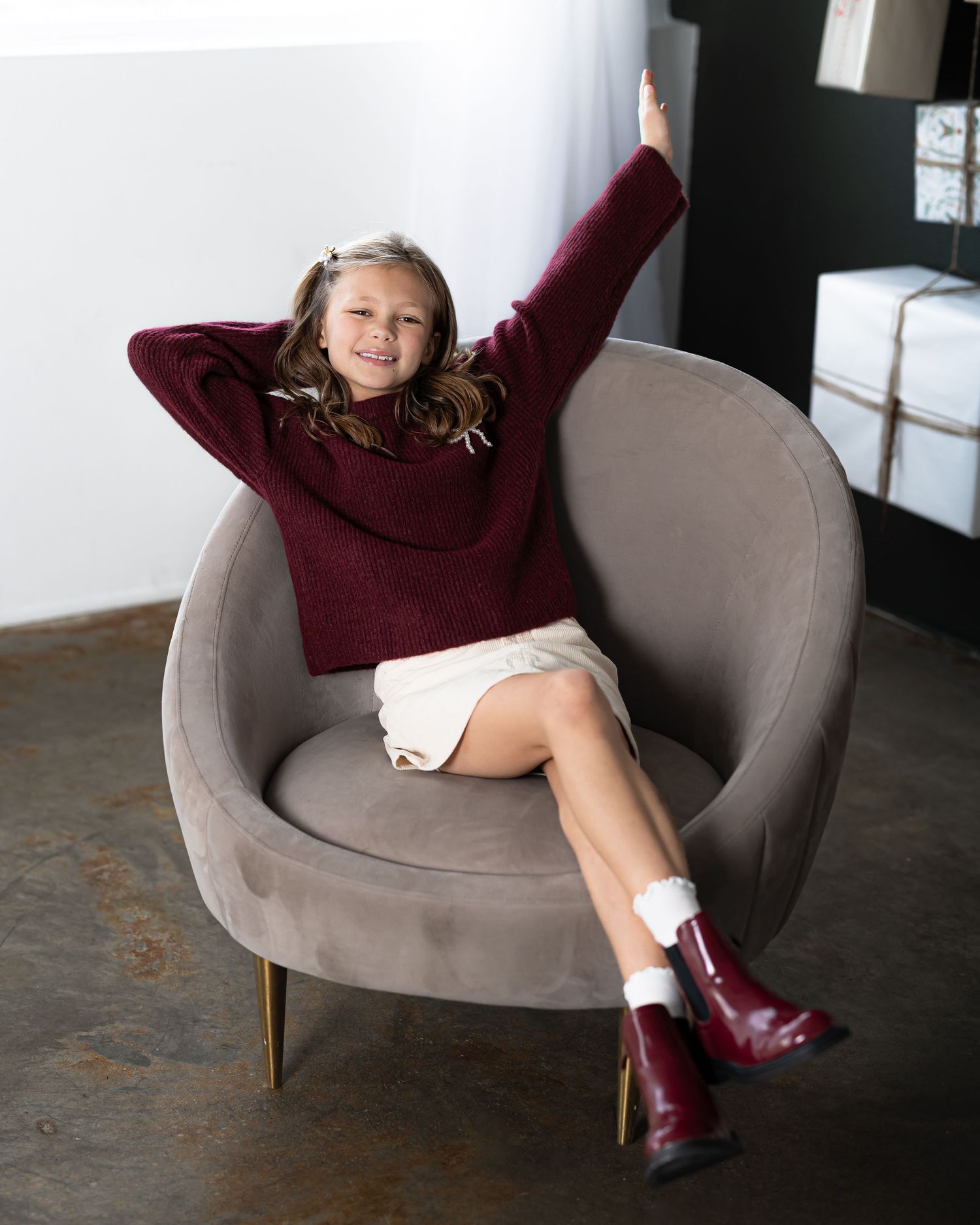 A girl in a burgundy sweater and boots lounges casually in a modern grey armchair against a dark background.