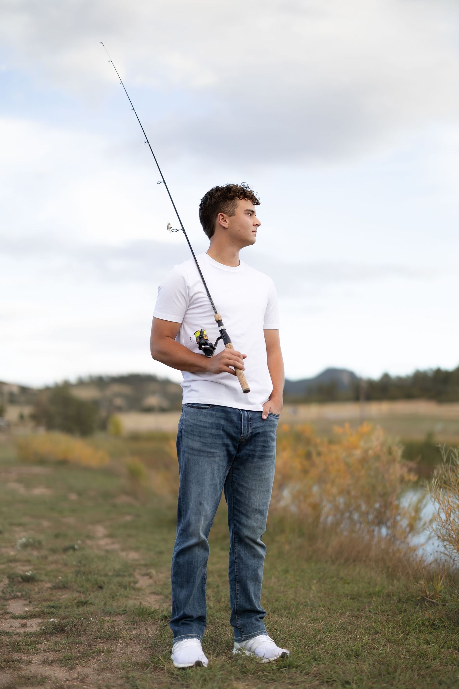 Person in white t-shirt and jeans holding fishing rod by lake during sunset.