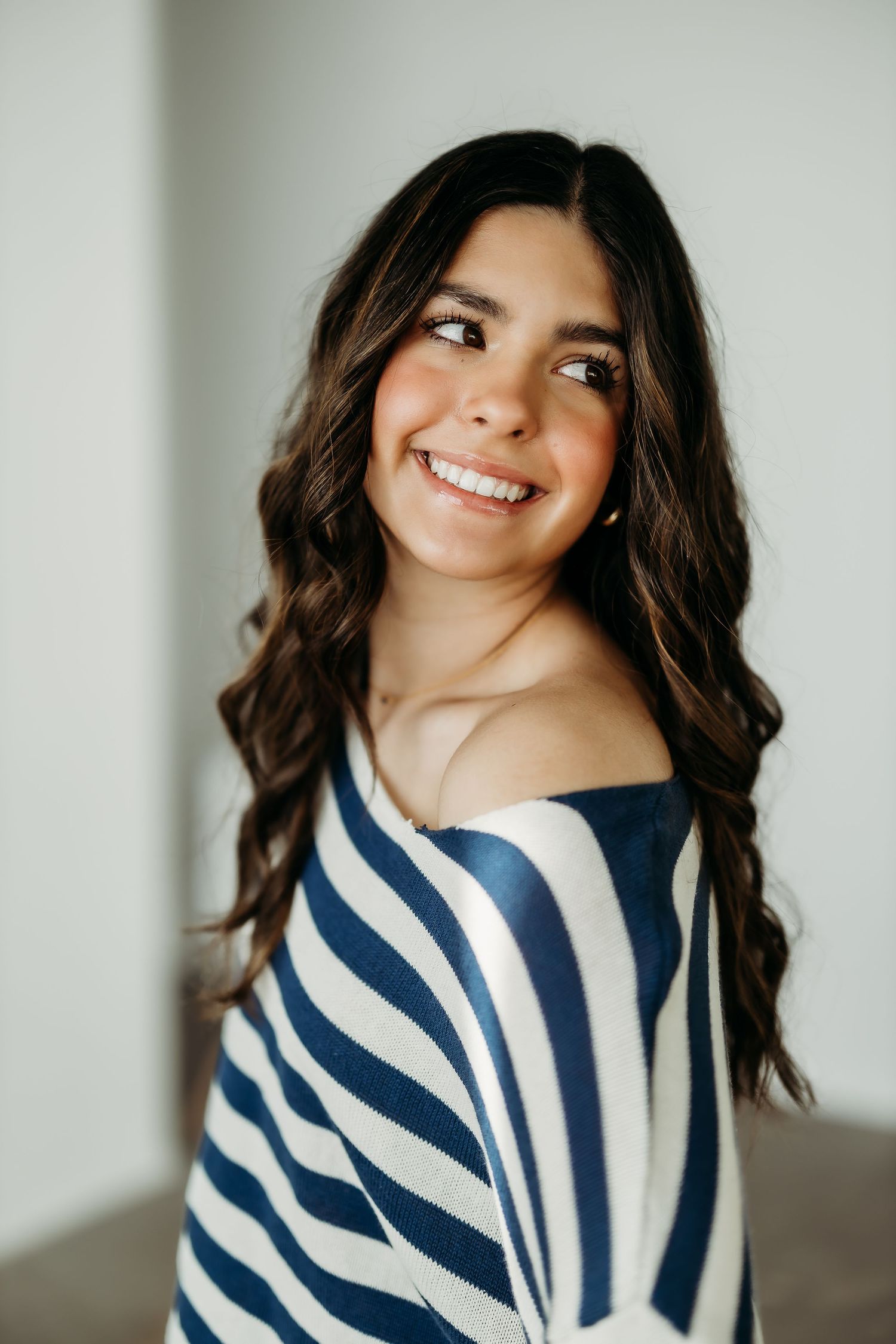 Beautiful 8th grade girl in a navy and white striped sweater poses against a light gray wall with natural lighting.