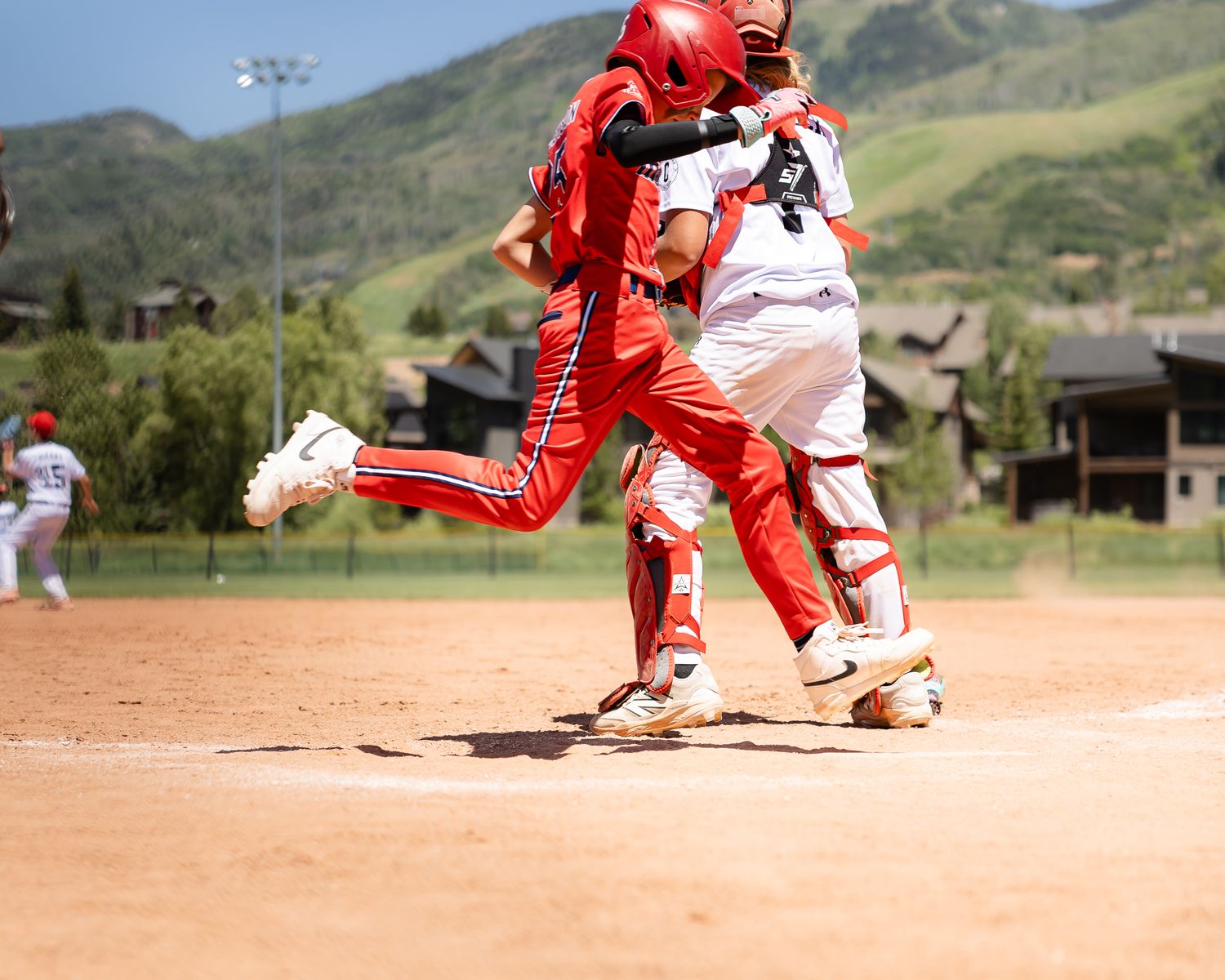 Baseball player in red uniform collides with catcher at home plate during a sunny outdoor game with mountains in background.