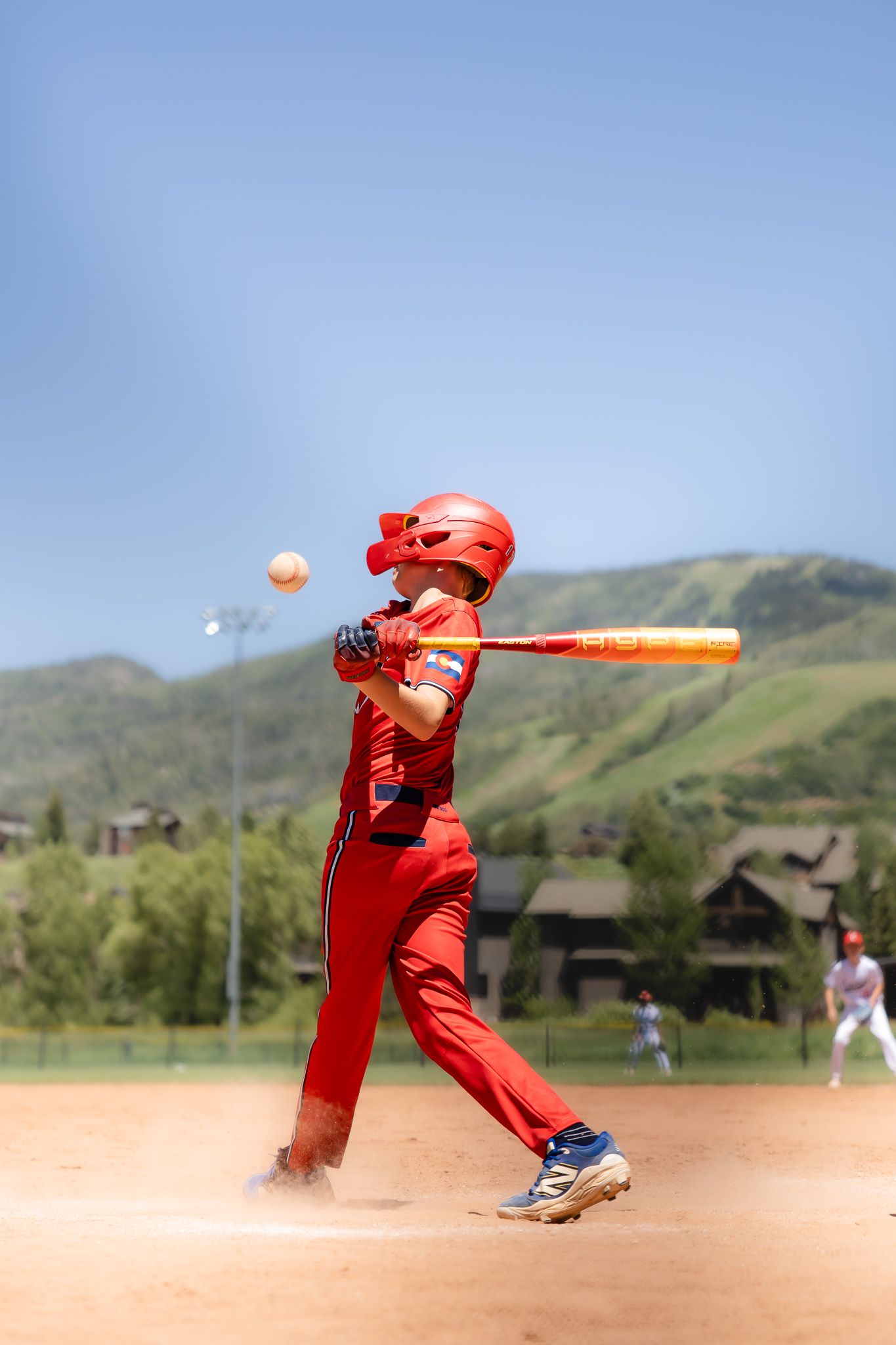Young baseball player in red uniform swinging bat at ball during a sunny outdoor game with green hills in background.