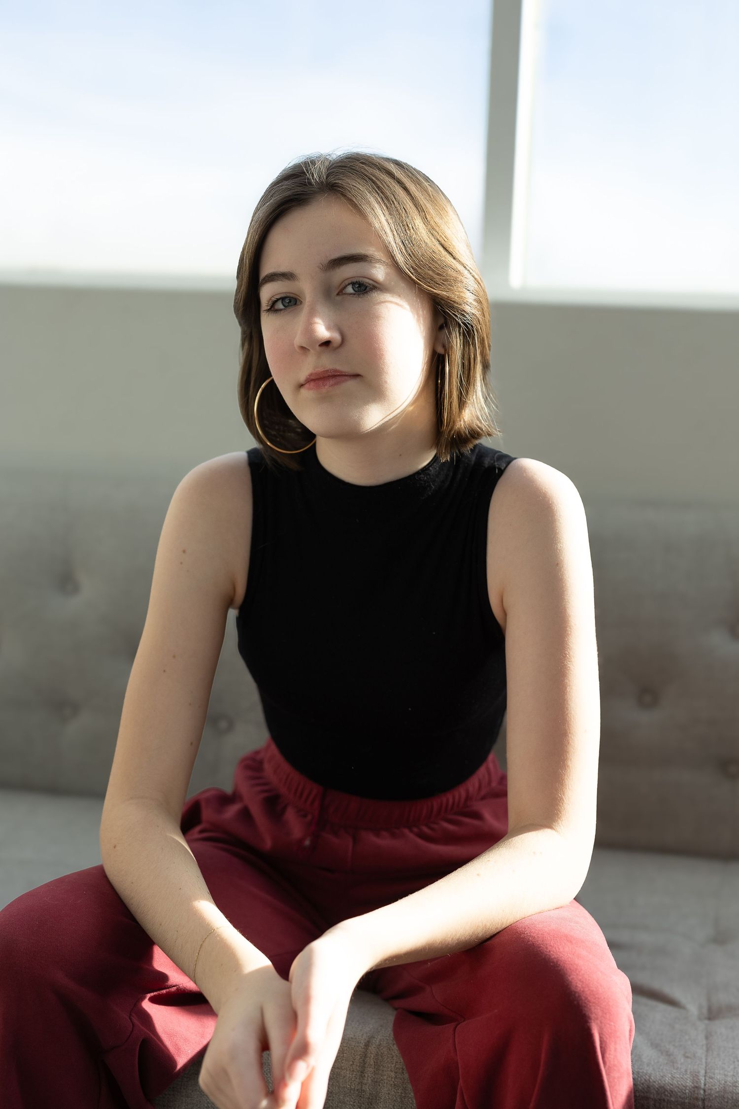 8th grade girl in black top and burgundy pants sits casually on grey couch in bright sunlit room.