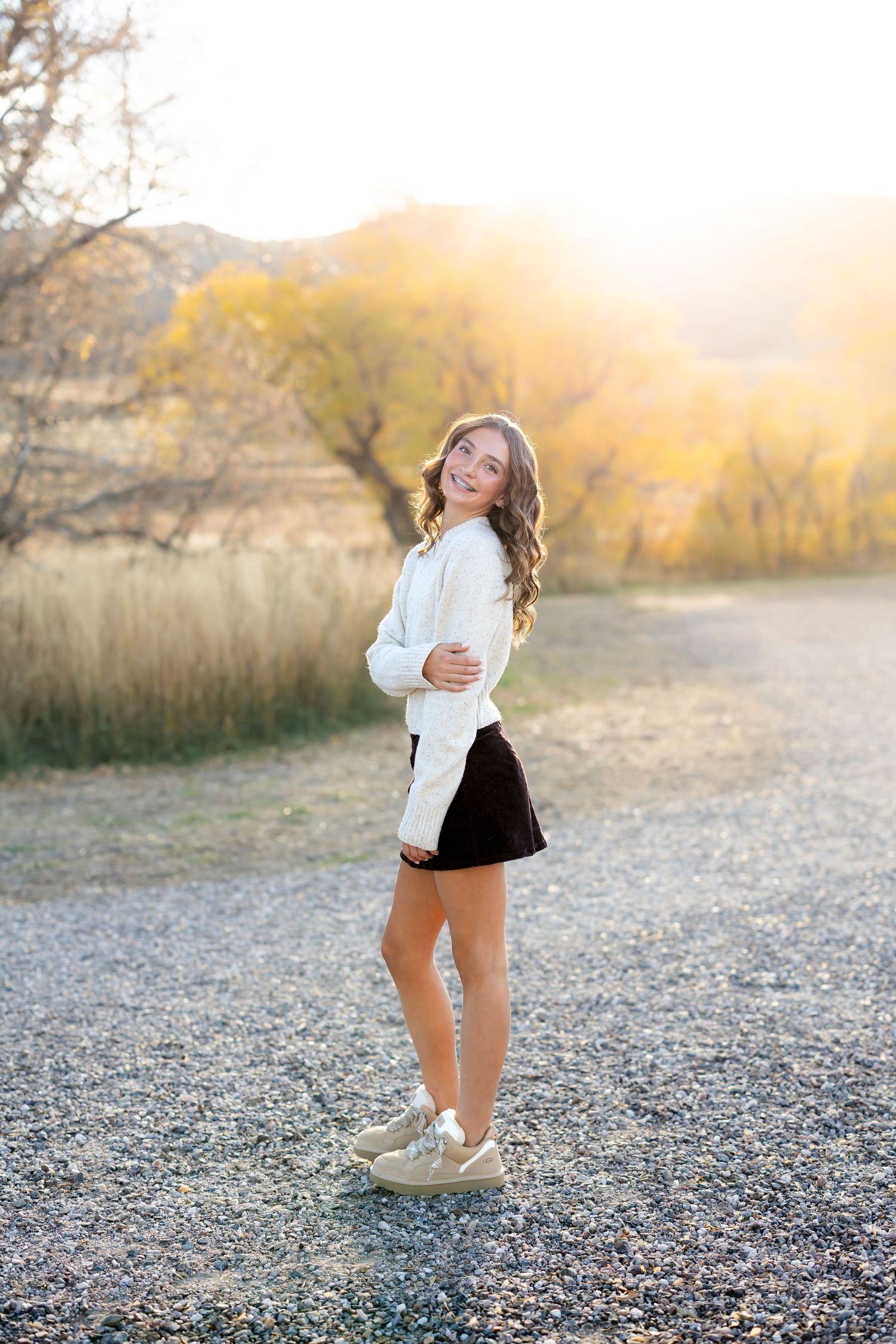 8th grade girl n golden sunlight showing someone in a white sweater and black skirt standing on gravel path near autumn trees.
