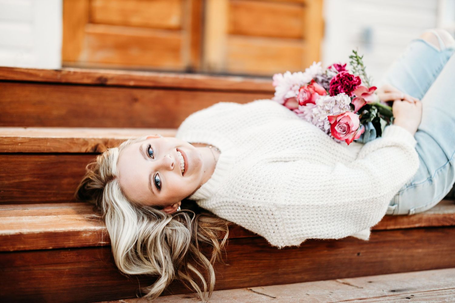 Person in white dress lying on wooden bench with flowers nearby.