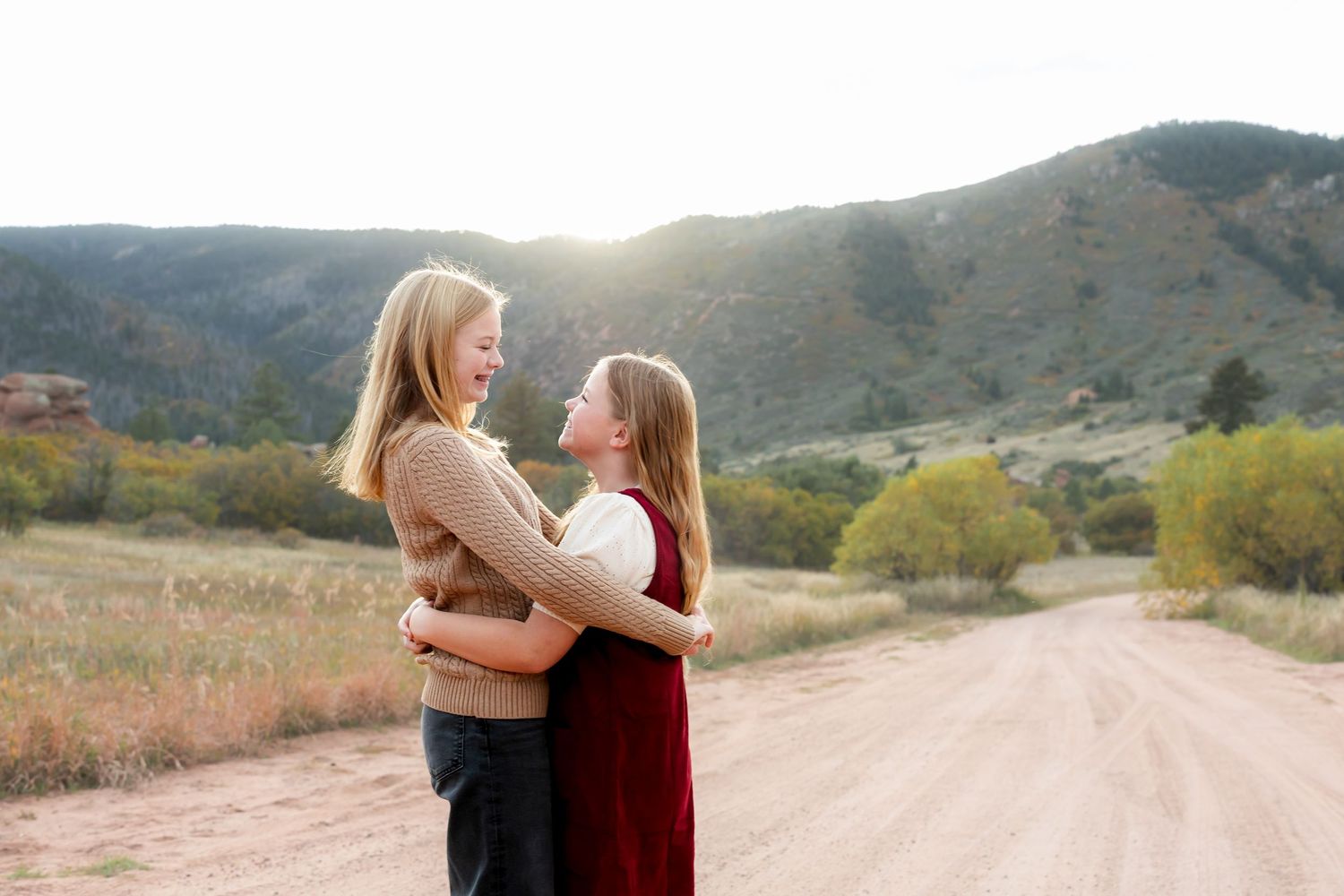 Two sisters embrace on a dirt road with rolling hills and mountains in the background at sunset.