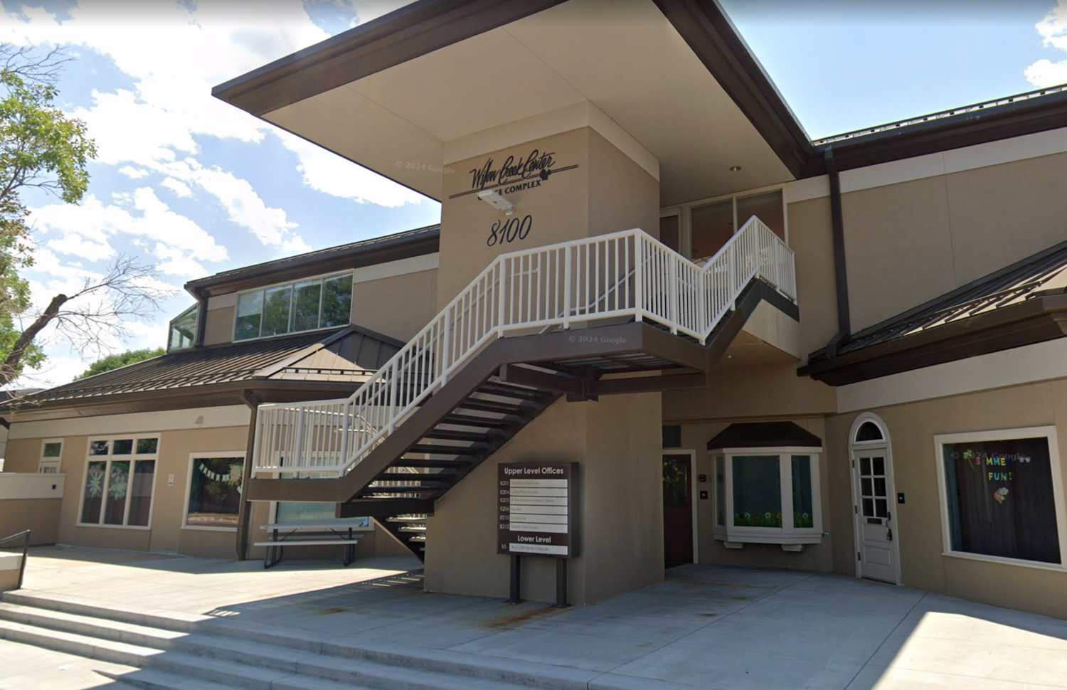Two-story beige commercial building with exterior staircase, white railings, and landscaped entrance on a sunny day.