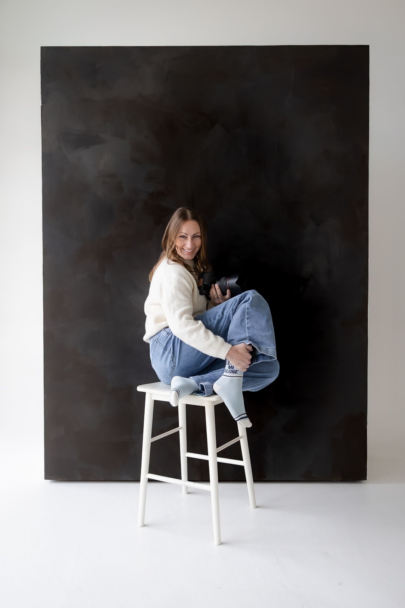 A person in a white sweater and jeans sits casually on a white bar stool against a dark backdrop in a studio setting.