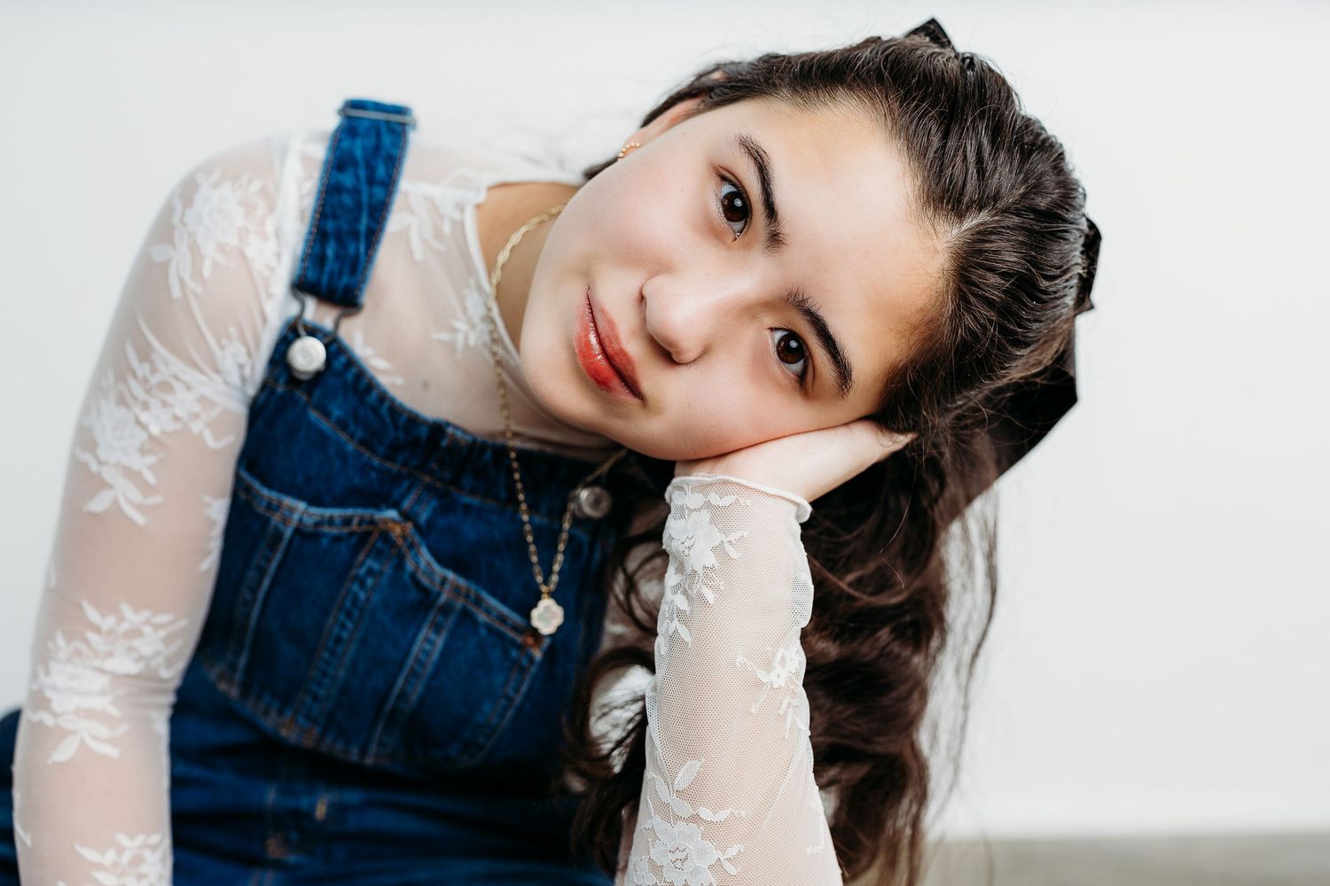 8th grade girl in denim overalls and white lace top poses playfully in a bright studio setting.