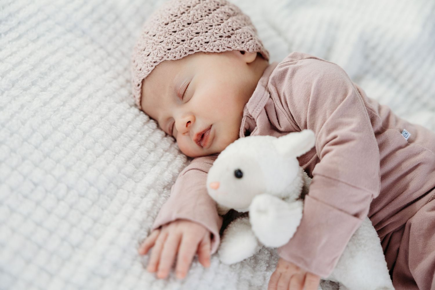 Baby sleeping on white bedding with plush bunny toy.