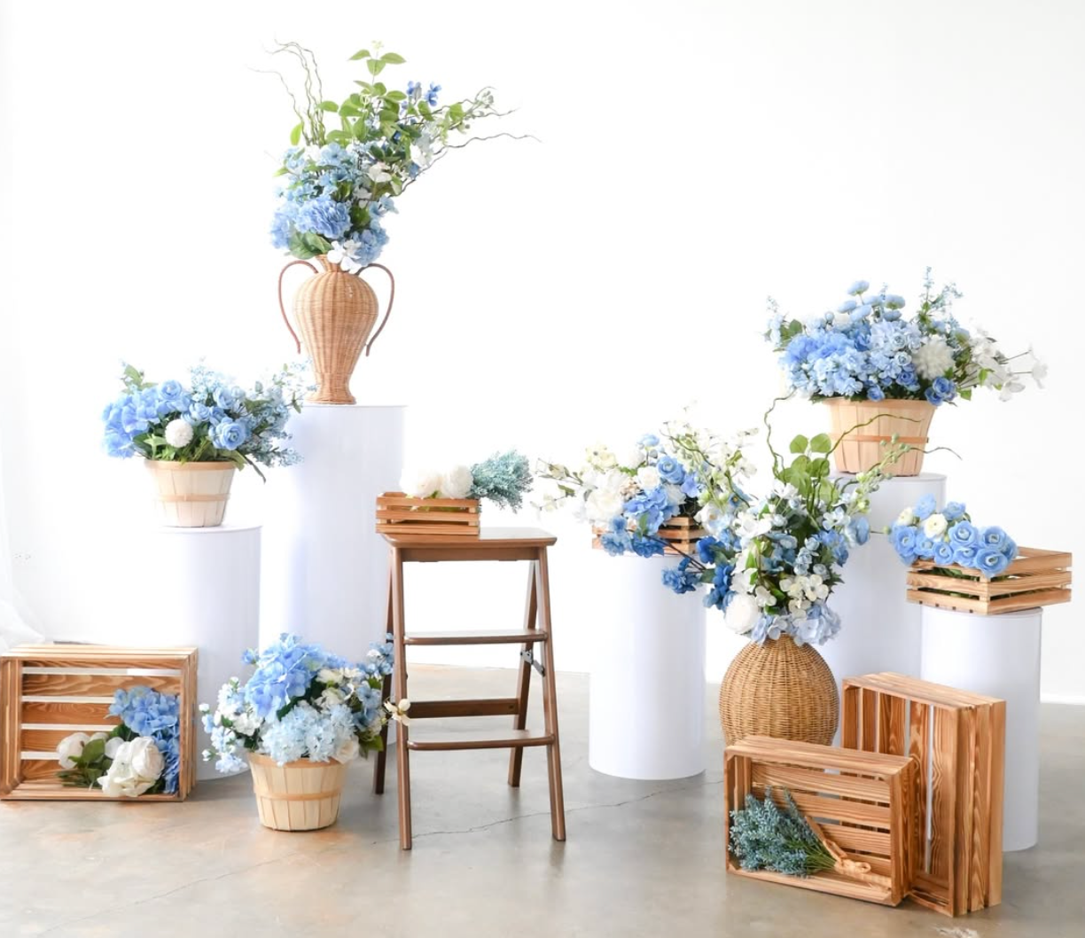 Blue hydrangea floral arrangements in wicker baskets displayed on white pedestals and wooden crates for an elegant event.