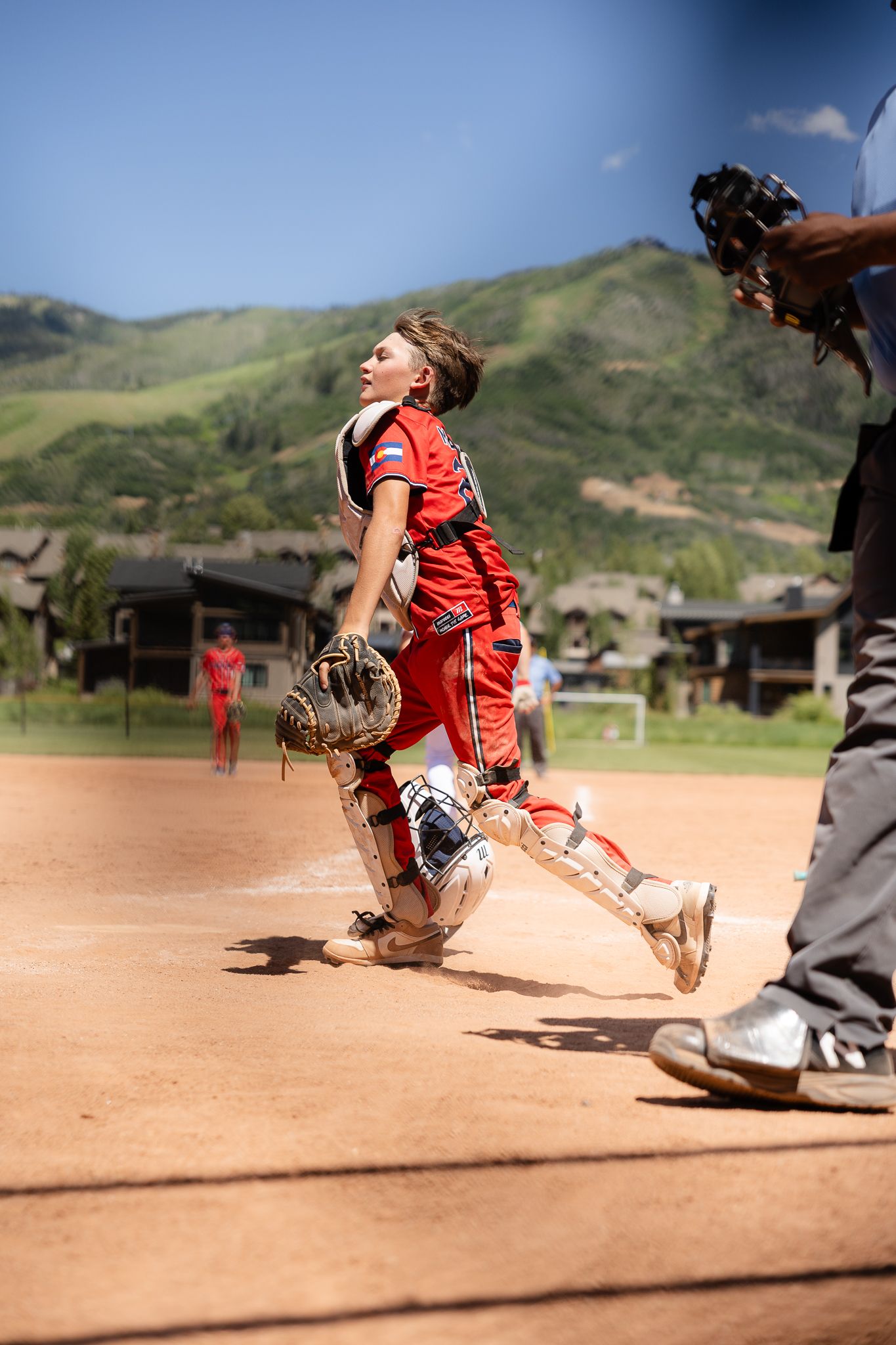 Young softball catcher in red uniform leaping to catch a pop fly during a sunny mountain backdrop game.