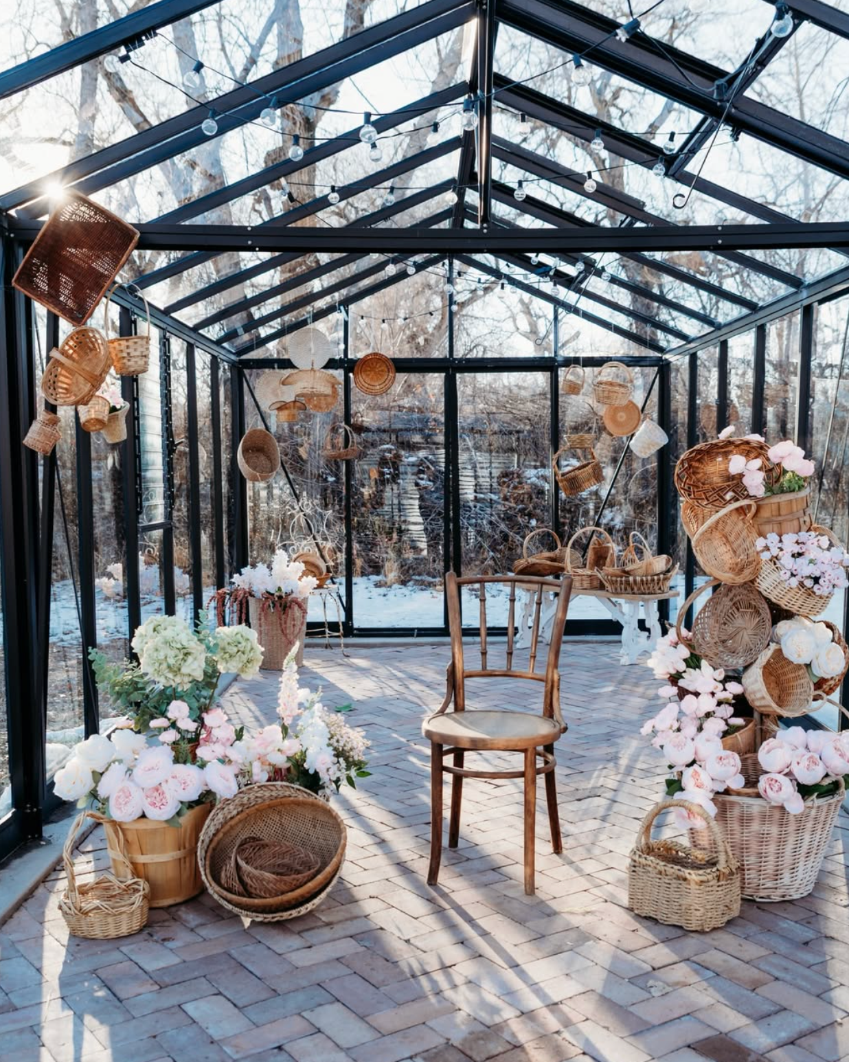 A sunlit glass greenhouse decorated with hanging wicker baskets and pink floral arrangements surrounding a wooden bentwood chair.