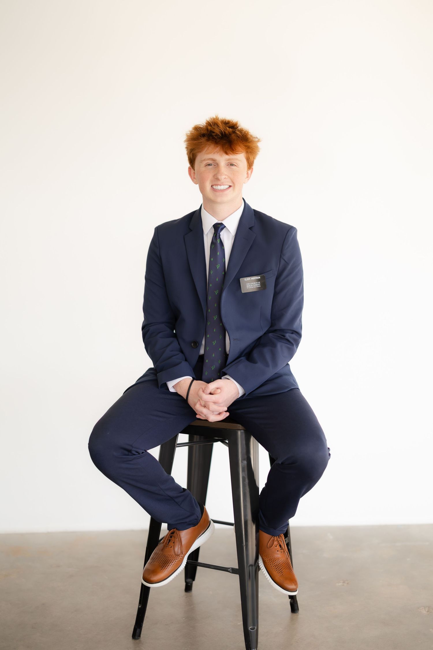 A person in a navy suit sits on a black stool against a white background in a professional portrait setting.