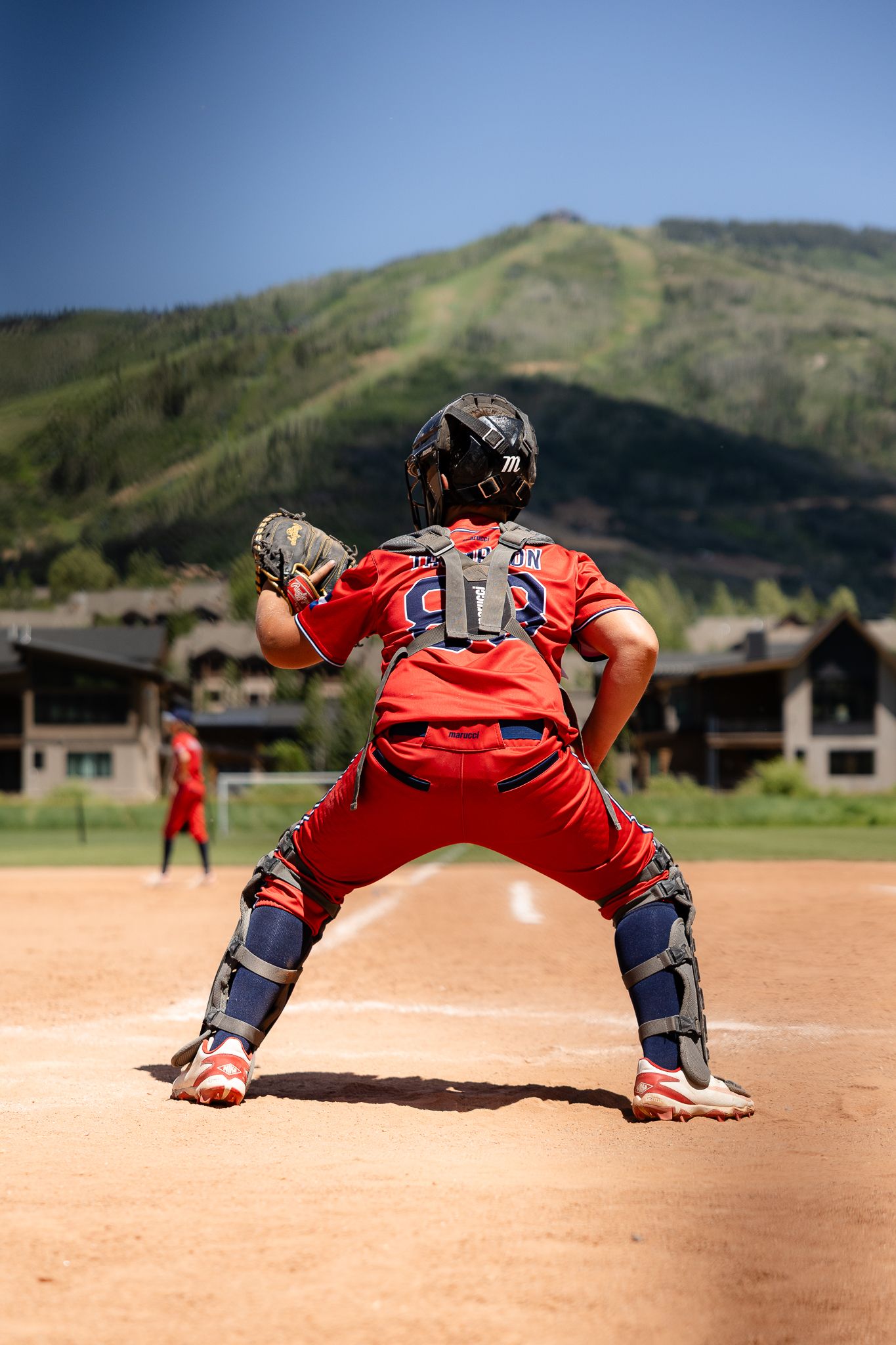Baseball catcher in red uniform crouching at home plate with mountain backdrop on sunny day.