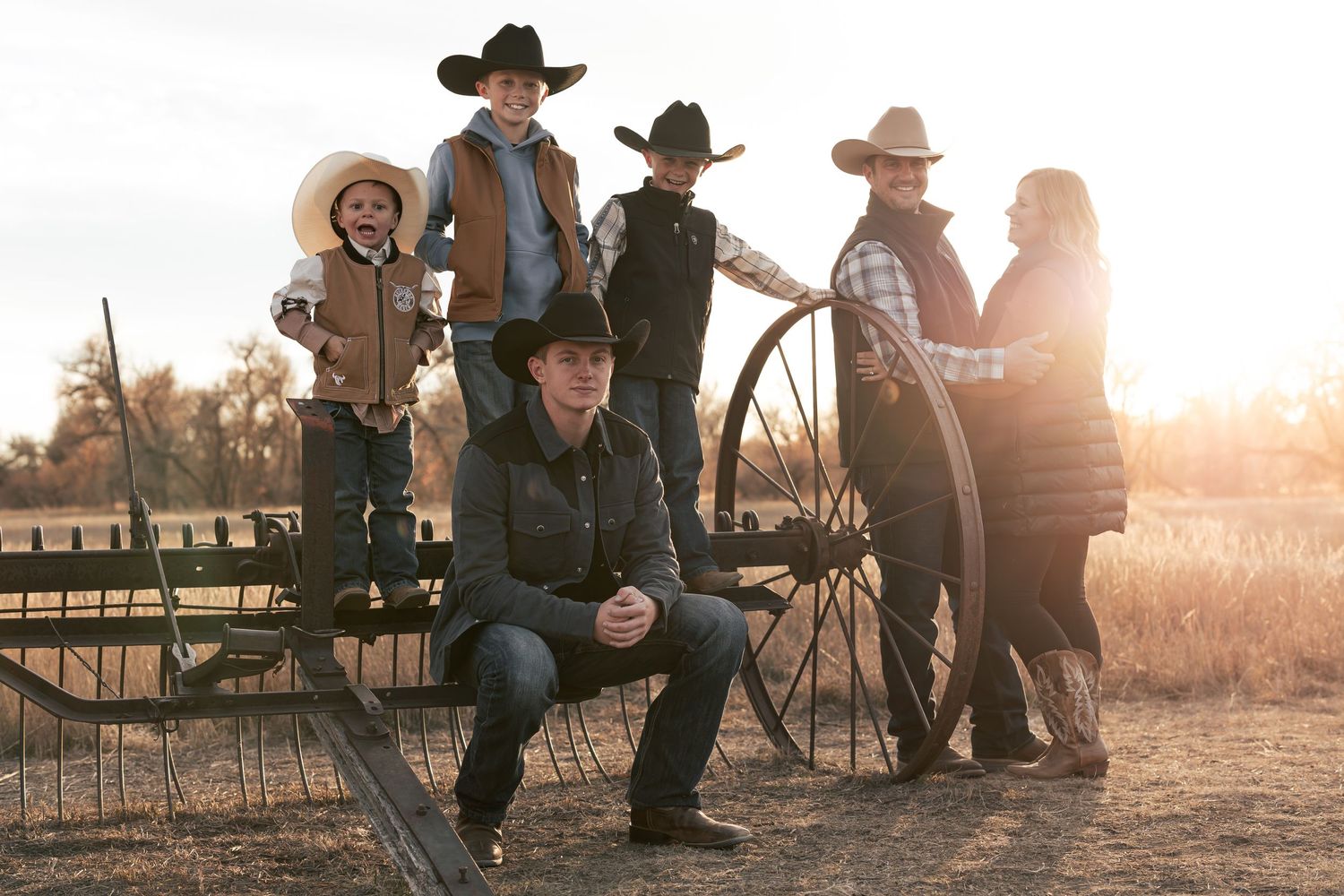 Group in western attire gathered around wagon wheel at sunset.