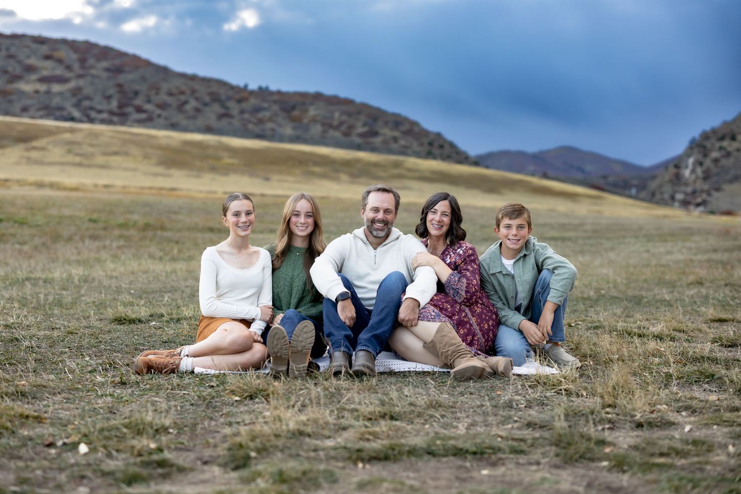 Group sits together on blanket in scenic mountain meadow with dog on sunny day.