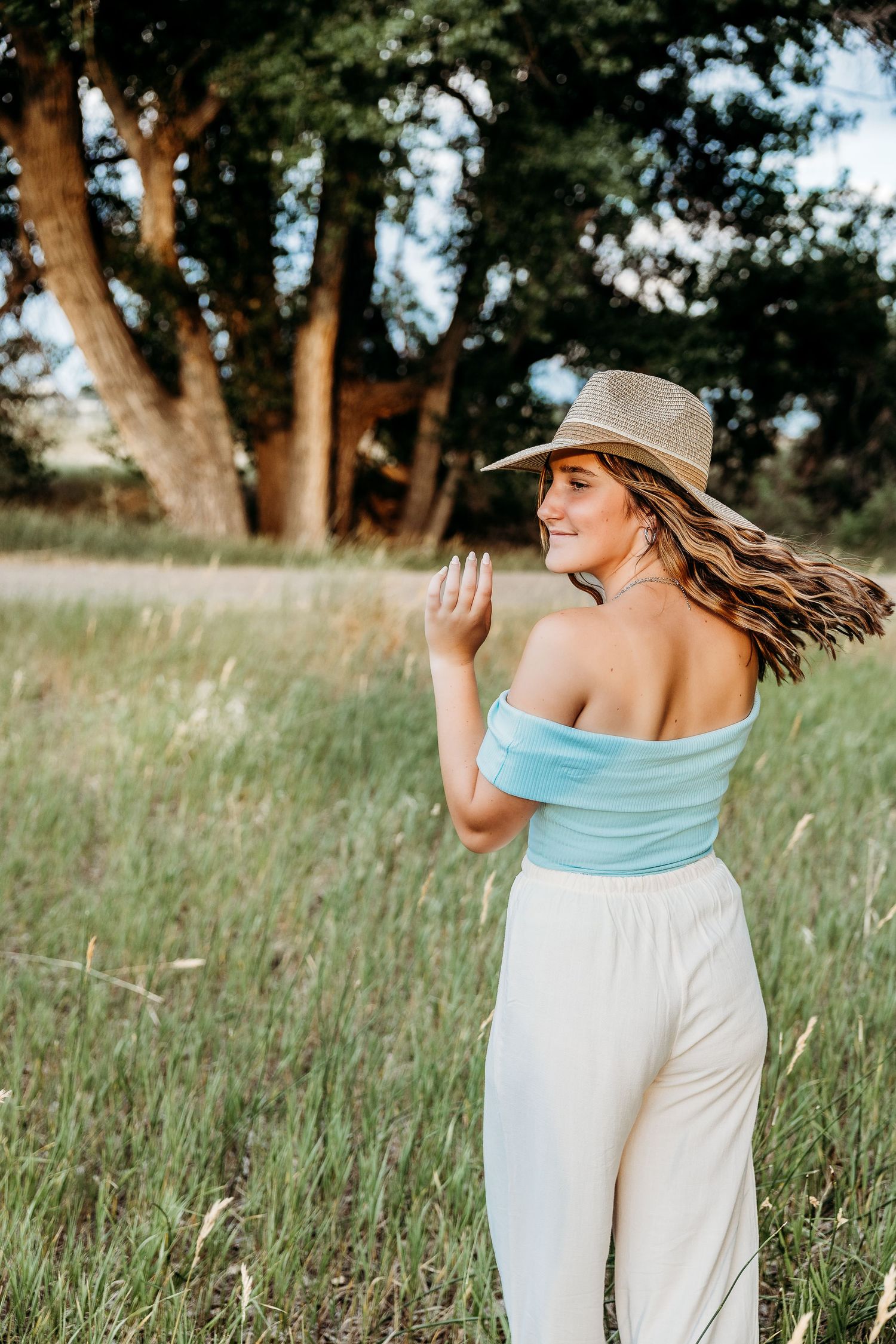 Woman in light blue top and white pants twirling gracefully in a sunny meadow with trees in background.