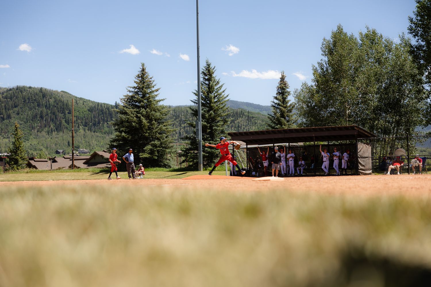 Youth baseball players in red uniforms play a game on a field surrounded by pine trees and mountains on a sunny day.