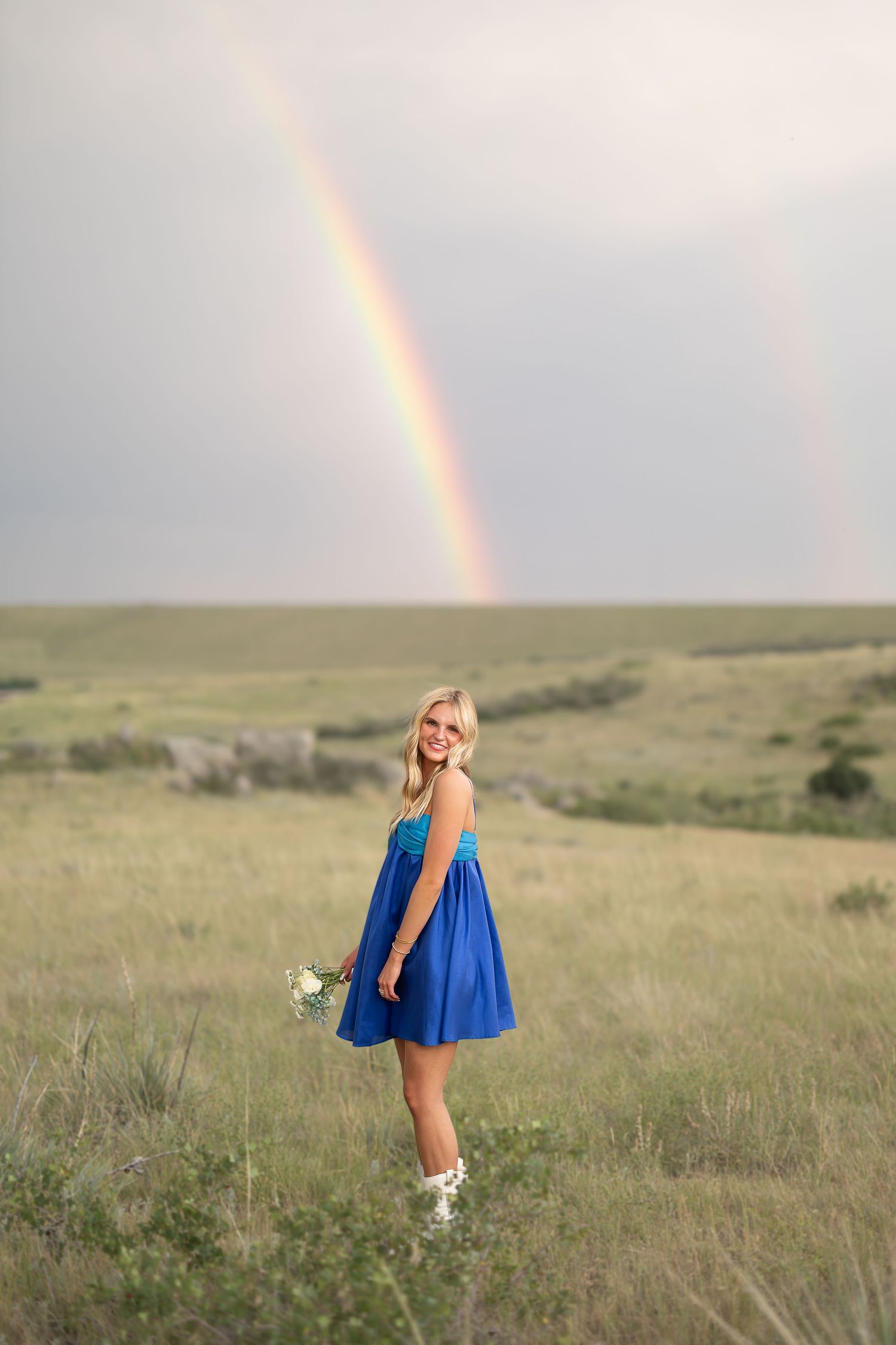 Person in blue dress twirling in field with rainbow visible in cloudy sky.